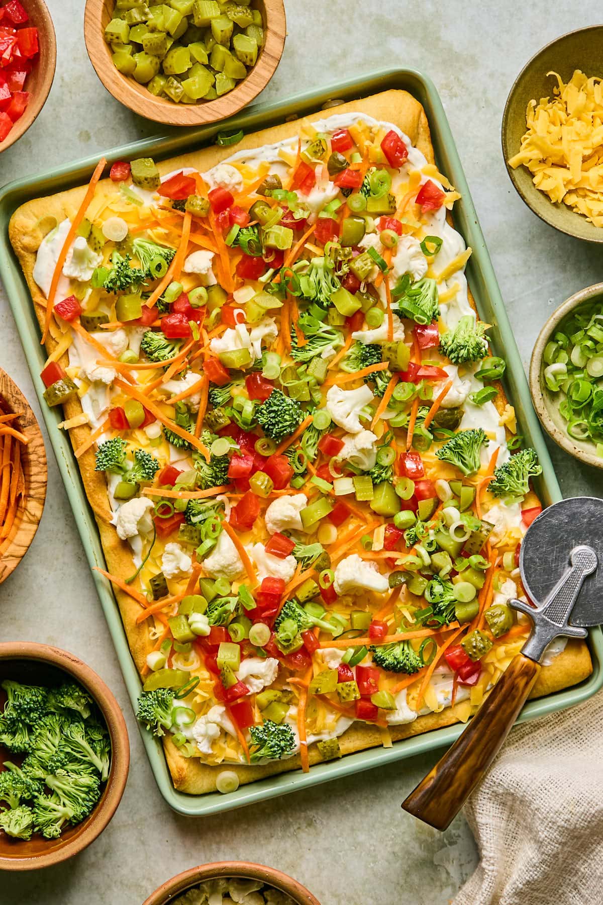 A colorful crescent roll vegetable pizza topped with broccoli, shredded carrots, diced tomatoes, grated cheese, and green onions on a rectangular baking sheet. A pizza cutter with a wooden handle rests nearby, surrounded by bowls of toppings.