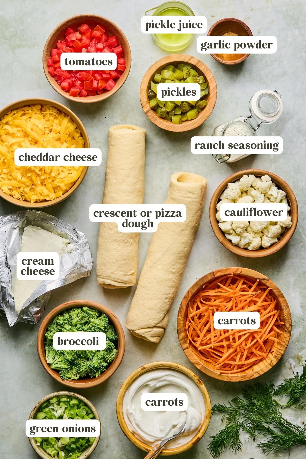 Assorted ingredients in bowls for a veggie pizza: tomatoes, cheddar cheese, crescent or pizza dough, broccoli, cream cheese, pickles, ranch seasoning, cauliflower, carrots, green onions, garlic powder, and pickle juice.
