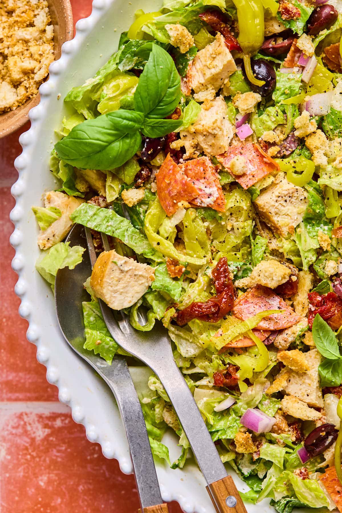 A close-up of a vibrant Italian chopped salad in a white dish. It features grilled chicken, lettuce, red onions, bell peppers, sun-dried tomatoes, black olives, and fresh basil. A serving spoon and fork rest in the dish. A small bowl of croutons is nearby.
