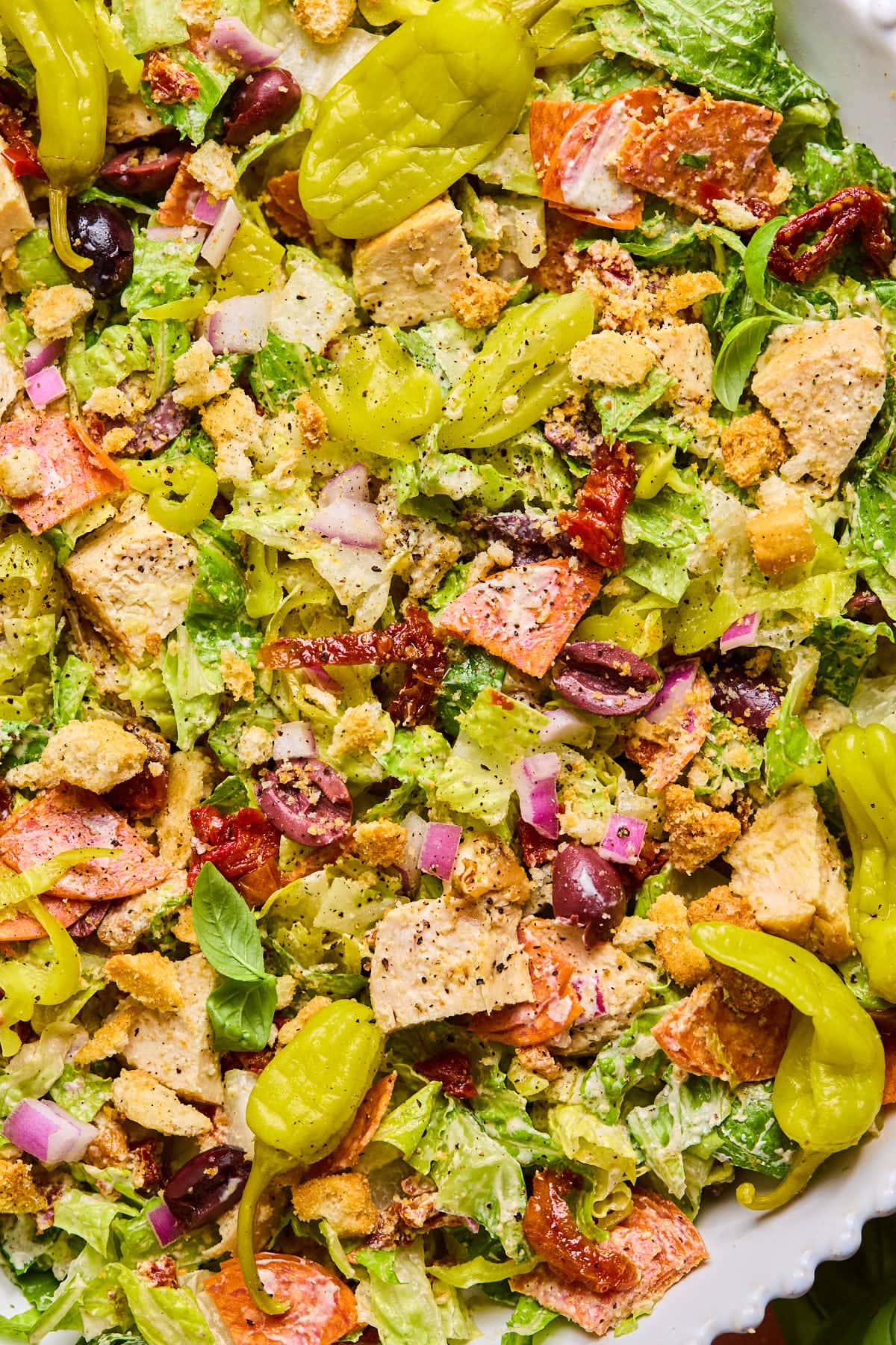 A vibrant Italian chopped salad featuring mixed greens, sliced pepperoncini, red onion, olives, sun-dried tomatoes, croutons, and chunks of grilled chicken. Everything is tossed together and seasoned with herbs and spices for a flavorful presentation.