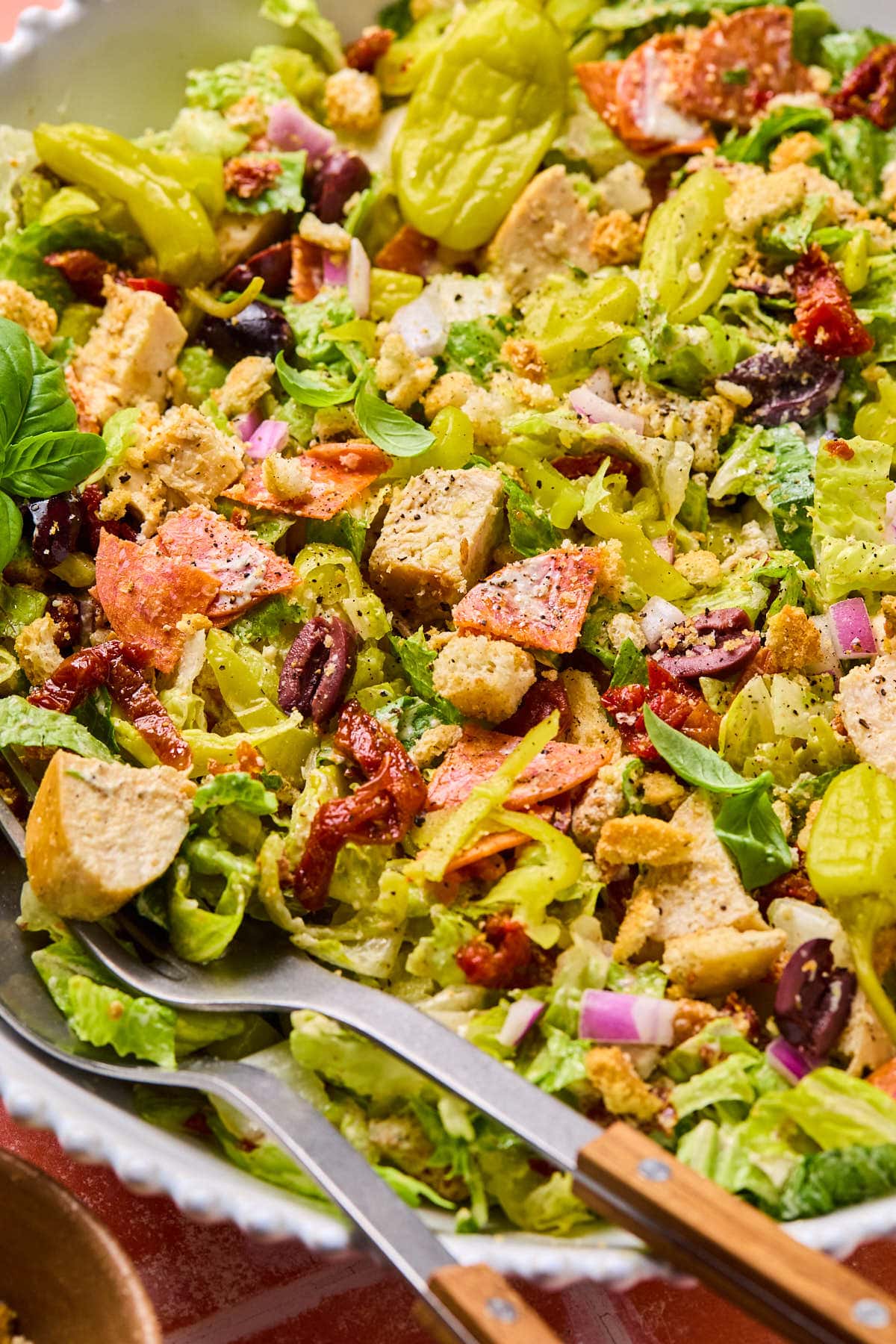 A close-up of a colorful Italian chopped salad in a bowl, featuring chopped lettuce, croutons, olives, red onions, pepperoncini, and sliced pepperoni. There are serving utensils with wooden handles resting in the salad.