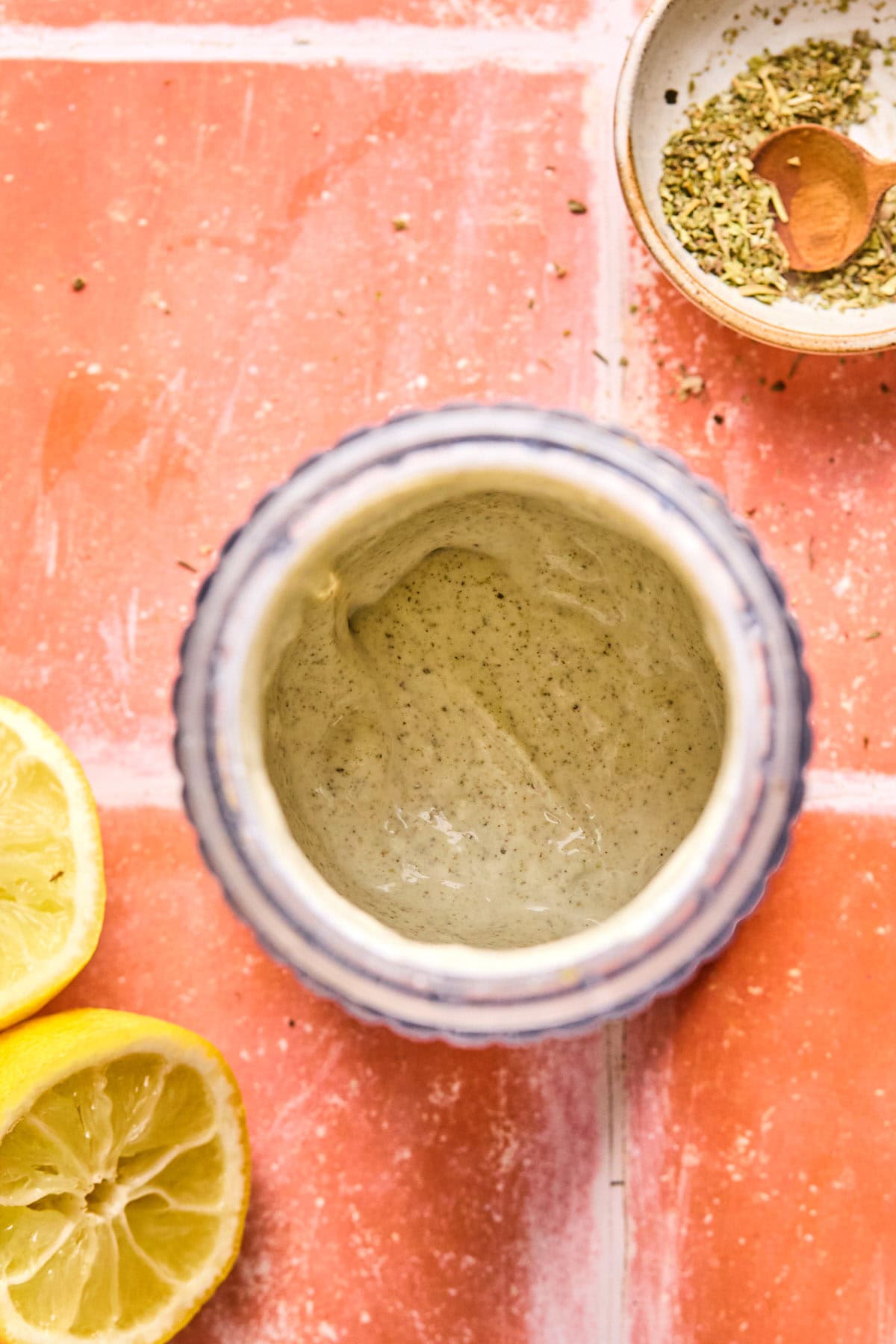 A jar of creamy Italian chopped salad dressing sits on a tiled surface, surrounded by partially sliced lemons and a small dish of herbs. The mixture inside the jar has a speckled texture, indicating spices or herbs blended in.