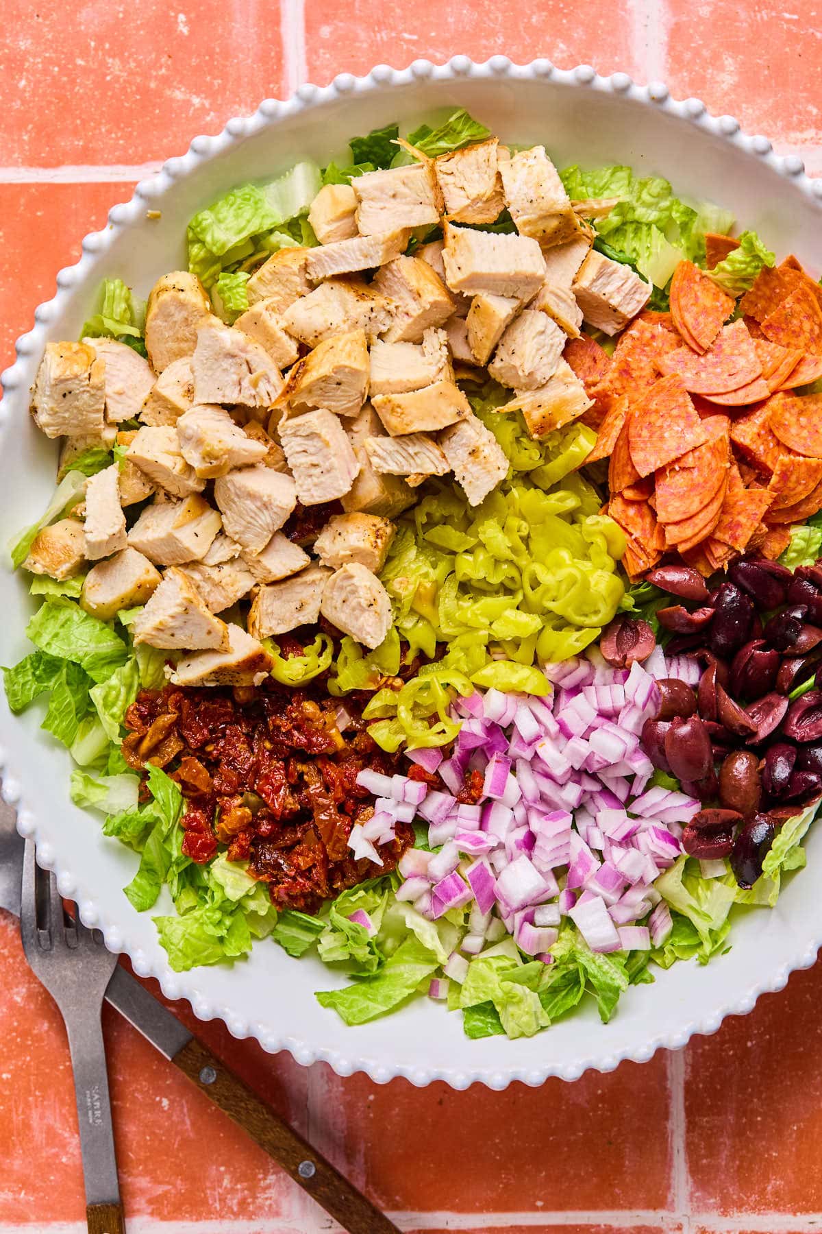 A vibrant salad in a white bowl featuring chopped lettuce topped with grilled chicken, pepperoni slices, banana peppers, sun-dried tomatoes, chopped red onions, and black olives. A fork rests beside the bowl on a red tiled surface.