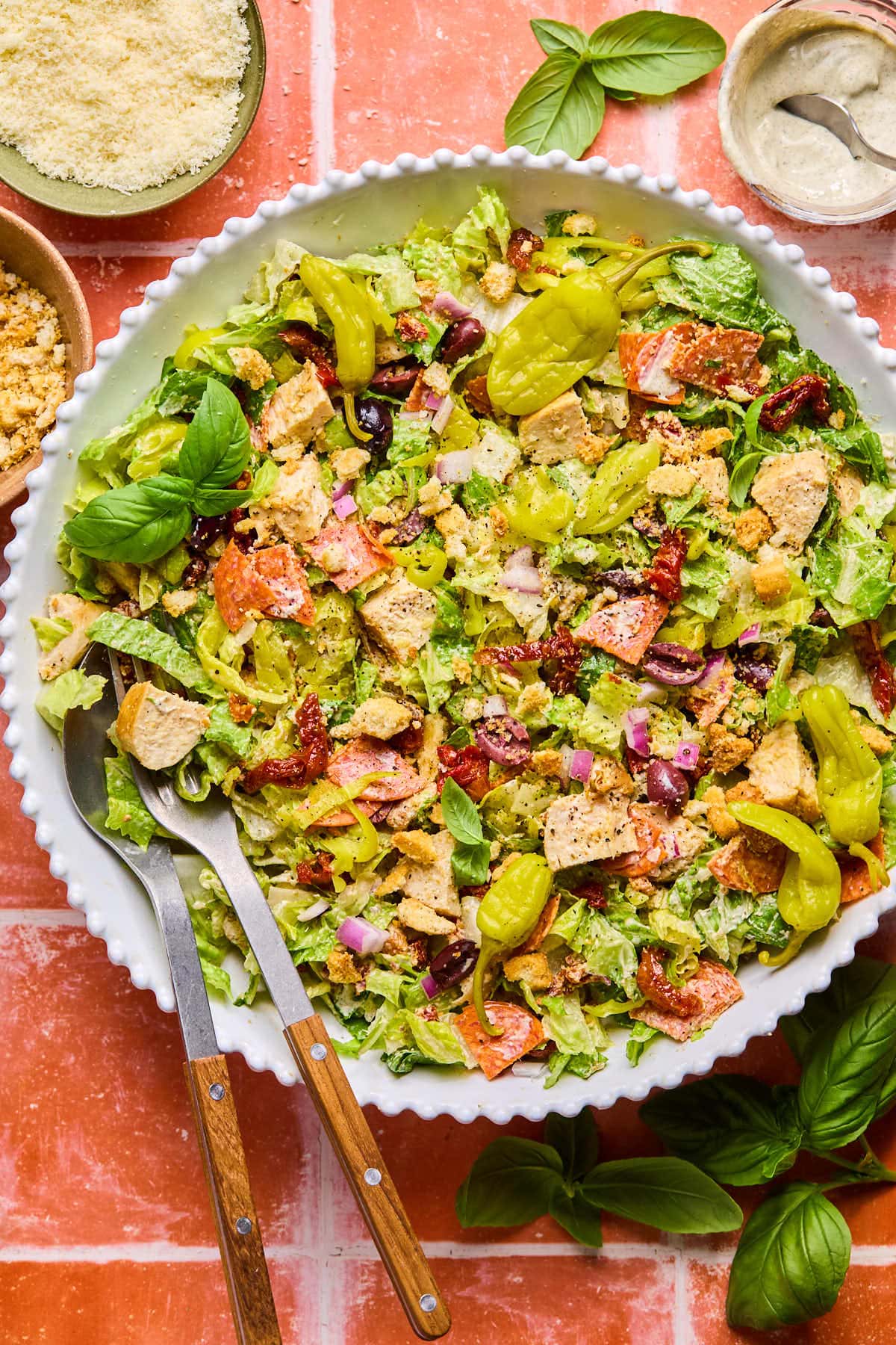 A colorful high protein Italian chopped salad in a white bowl on a red tile surface, featuring chopped lettuce, croutons, sliced tomatoes, red onions, olives, and pepperoncini, garnished with fresh basil. A bowl of grated cheese and a creamy dressing are on the side.