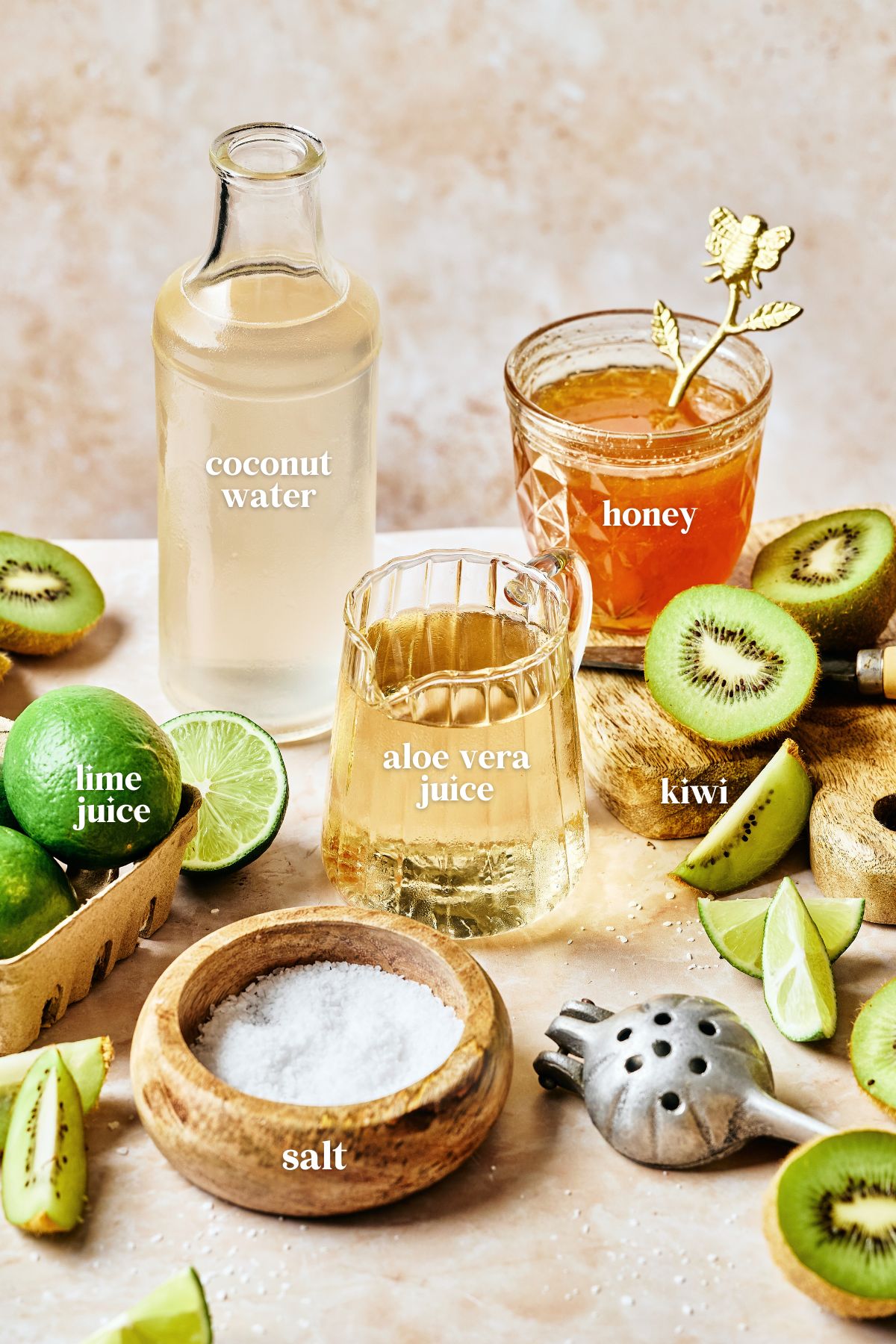 A refreshing drink setup featuring a bottle of coconut water, a honey jar with a decorative stirrer, and a glass of aloe vera juice. Surrounded by sliced kiwis, limes, and a wooden bowl of salt on a light textured surface.