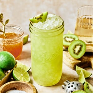 A tall glass filled with a bright green, icy kiwi homemade gatorade is garnished with a kiwi slice. Surrounding it are sliced kiwis, lime halves, a small bowl of salt, a honey jar, and a juicer on a beige surface.