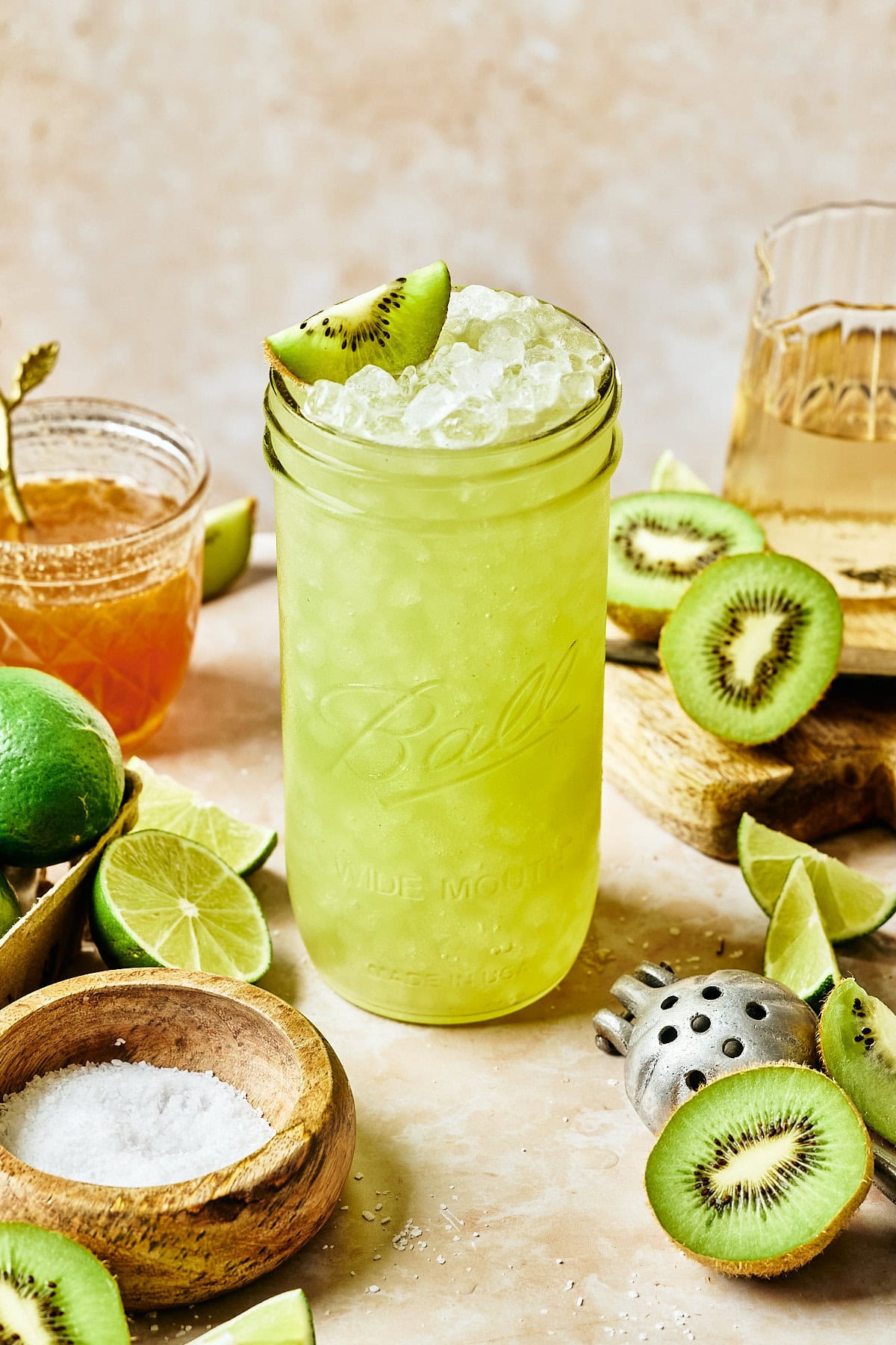 A tall glass filled with a bright green, icy kiwi homemade gatorade is garnished with a kiwi slice. Surrounding it are sliced kiwis, lime halves, a small bowl of salt, a honey jar, and a juicer on a beige surface.