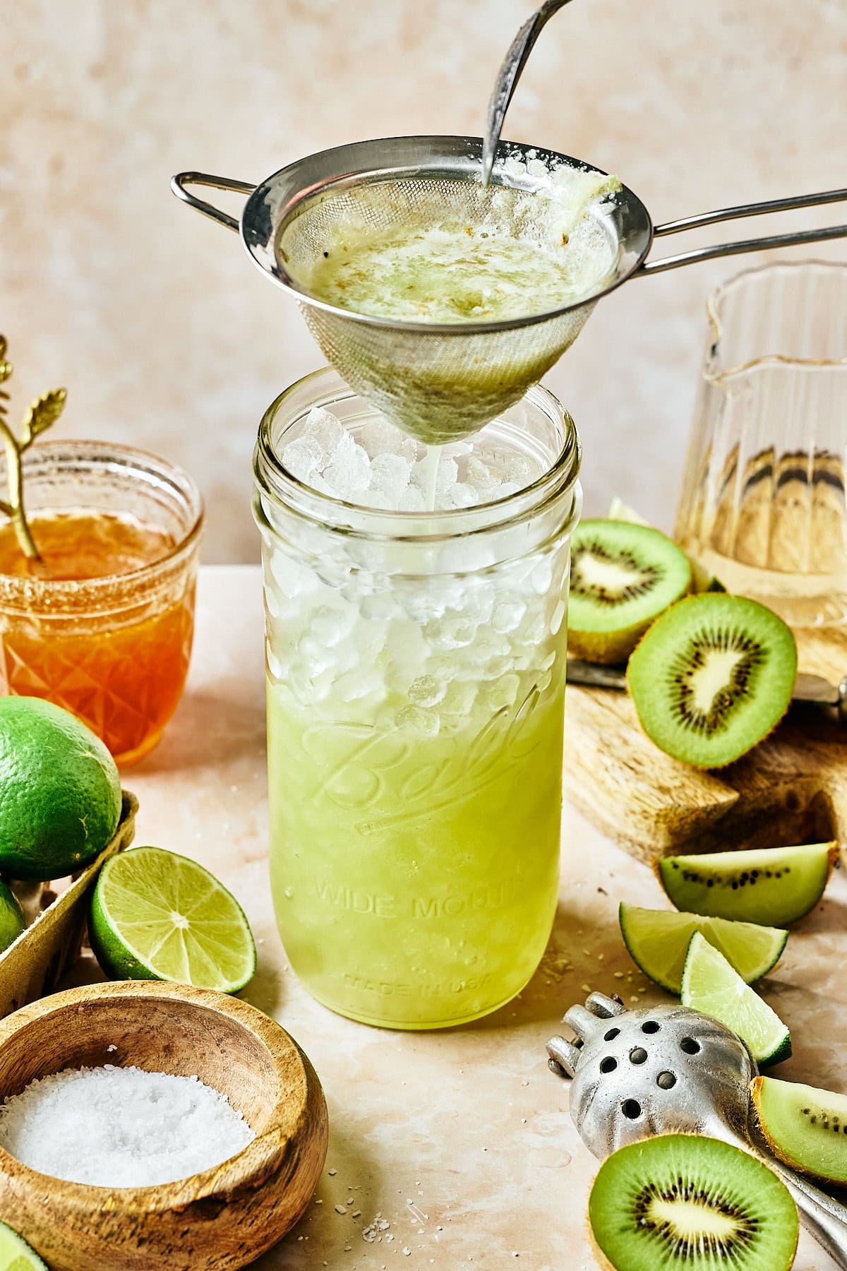 A glass jar filled with crushed ice and green liquid is being strained into. Surrounding it are kiwi slices, lime wedges, honey in a jar, and a bowl of salt. A citrus juicer and a metal strainer are also visible on the light-colored surface.