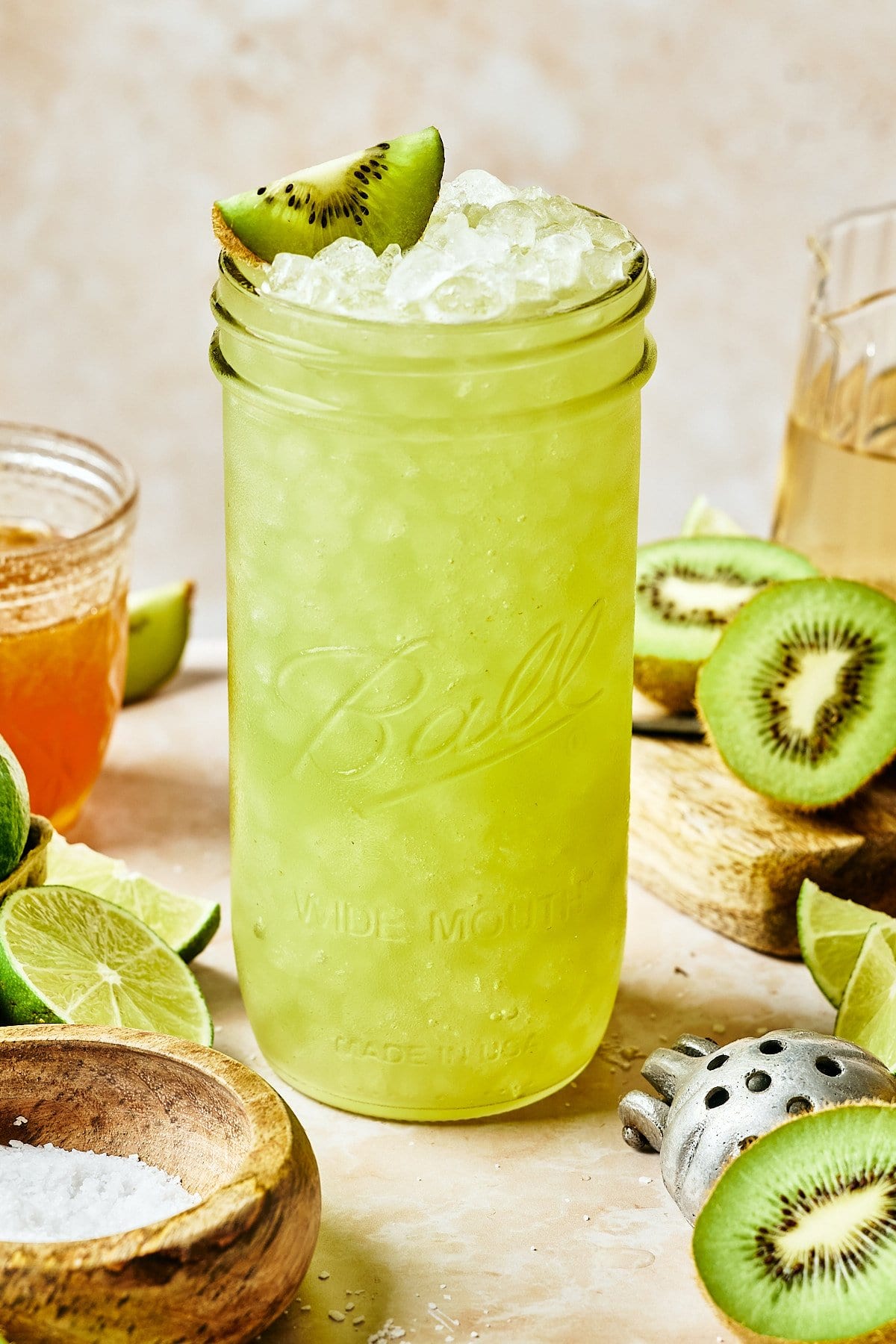 A refreshing homemade gatorade drink in a Ball mason jar, topped with crushed ice and a kiwi slice. Surrounded by fresh kiwi halves, lime wedges, a glass of liquid, a bowl of honey, and a salt holder, all on a light-colored surface.
