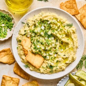 dill pickle hummus topped with crunchy chopped pickles and sprigs of dill, with three pita chips resting atop.
