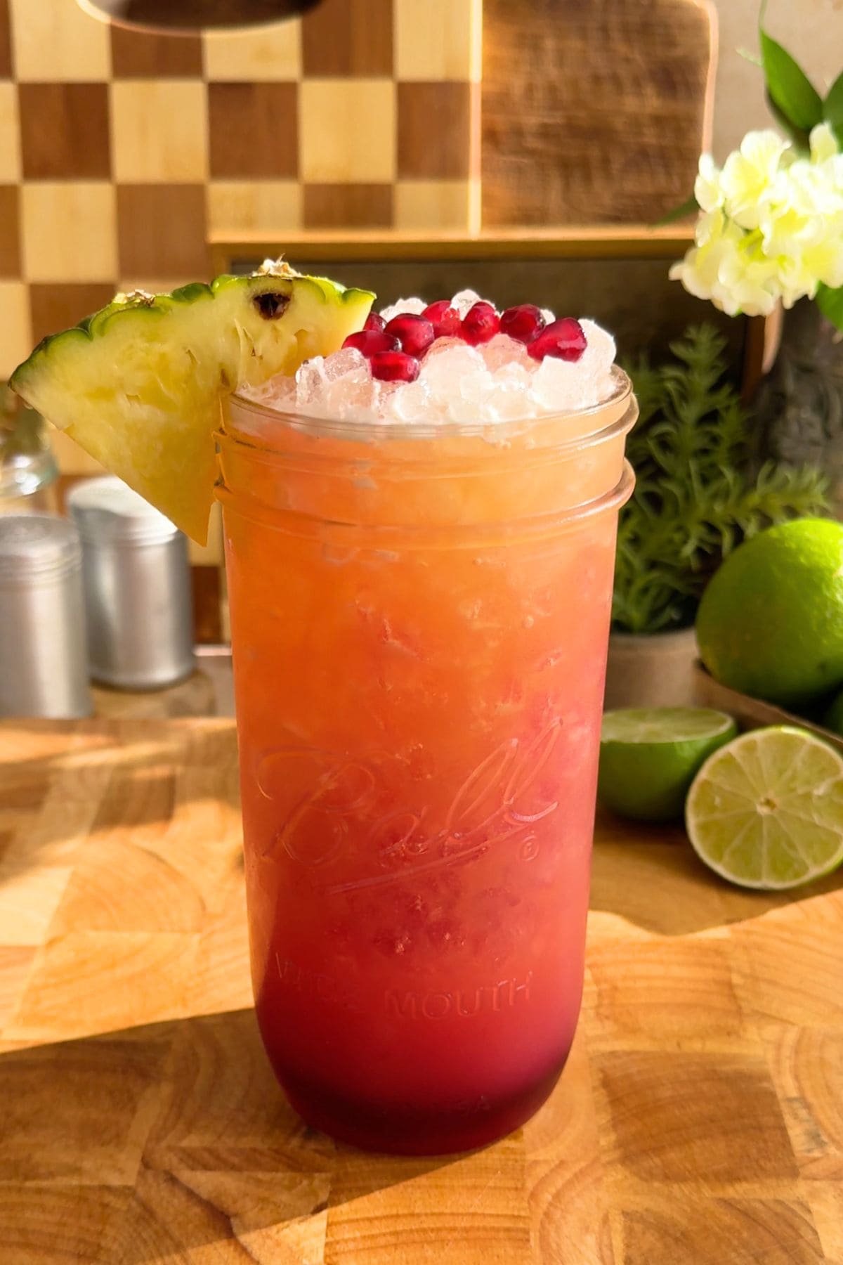A vibrant glass of pineapple pomegranate Gatorade with a layered golden yellow and ruby red color, served over ice with fresh fruit garnishes. Behind the glass are decorative plants, flowers, and fresh limes all in front of a wooden checkerboard surface.