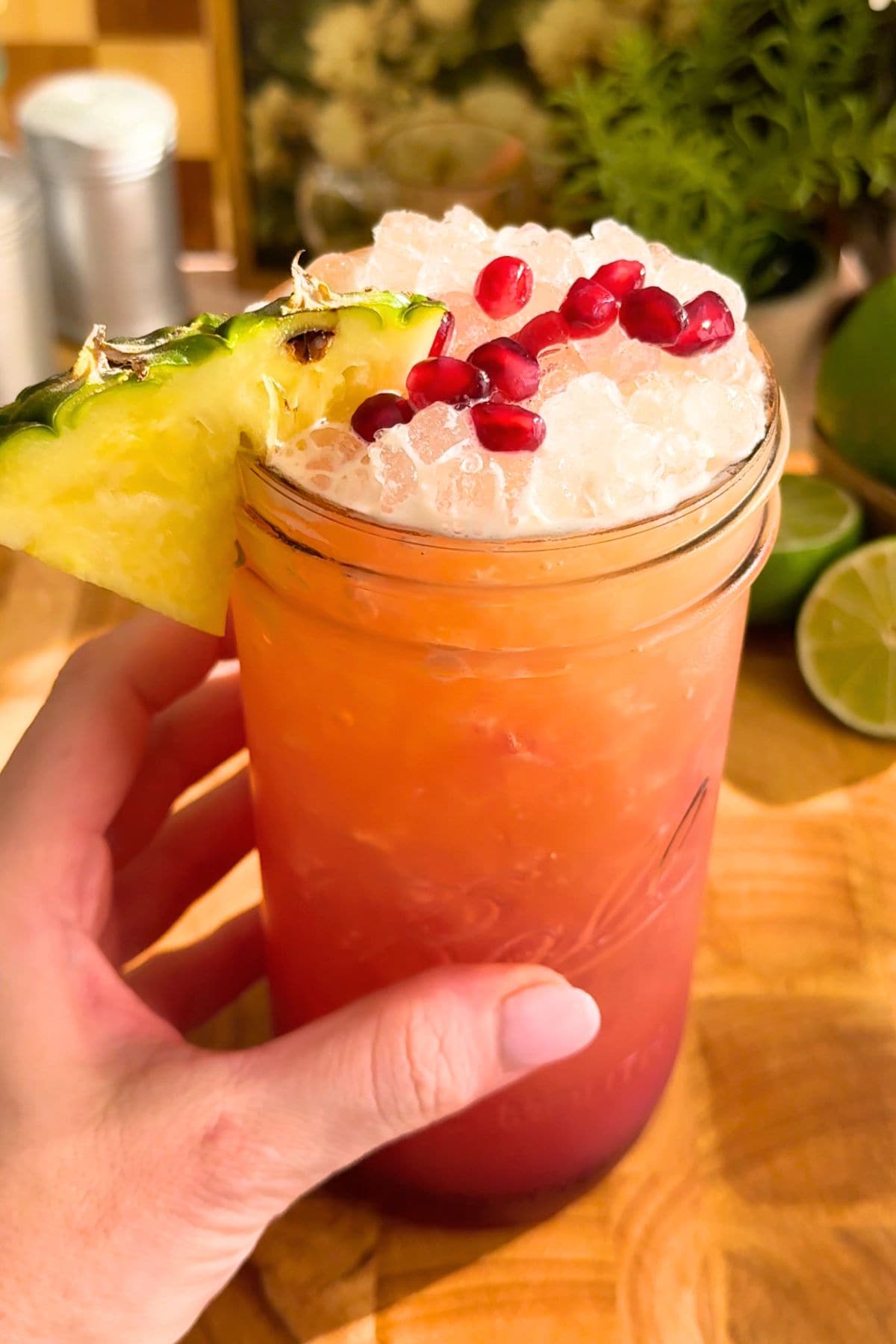 Close-up of a hand holding a homemade pineapple pomegranate Gatorade garnished with pomegranate arils and a pineapple wedge. Behind the glass is decorative plants and fresh lime halves.