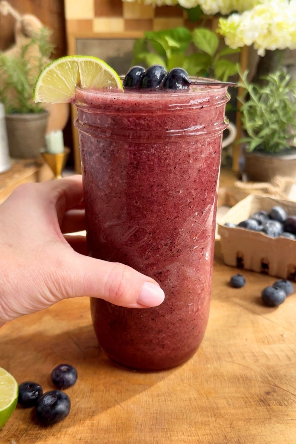 A hand holding a glass of homemade blueberry Gatorade with lime garnish, and plump blueberries on a wooden surface with decorative plants and blueberries behind it.