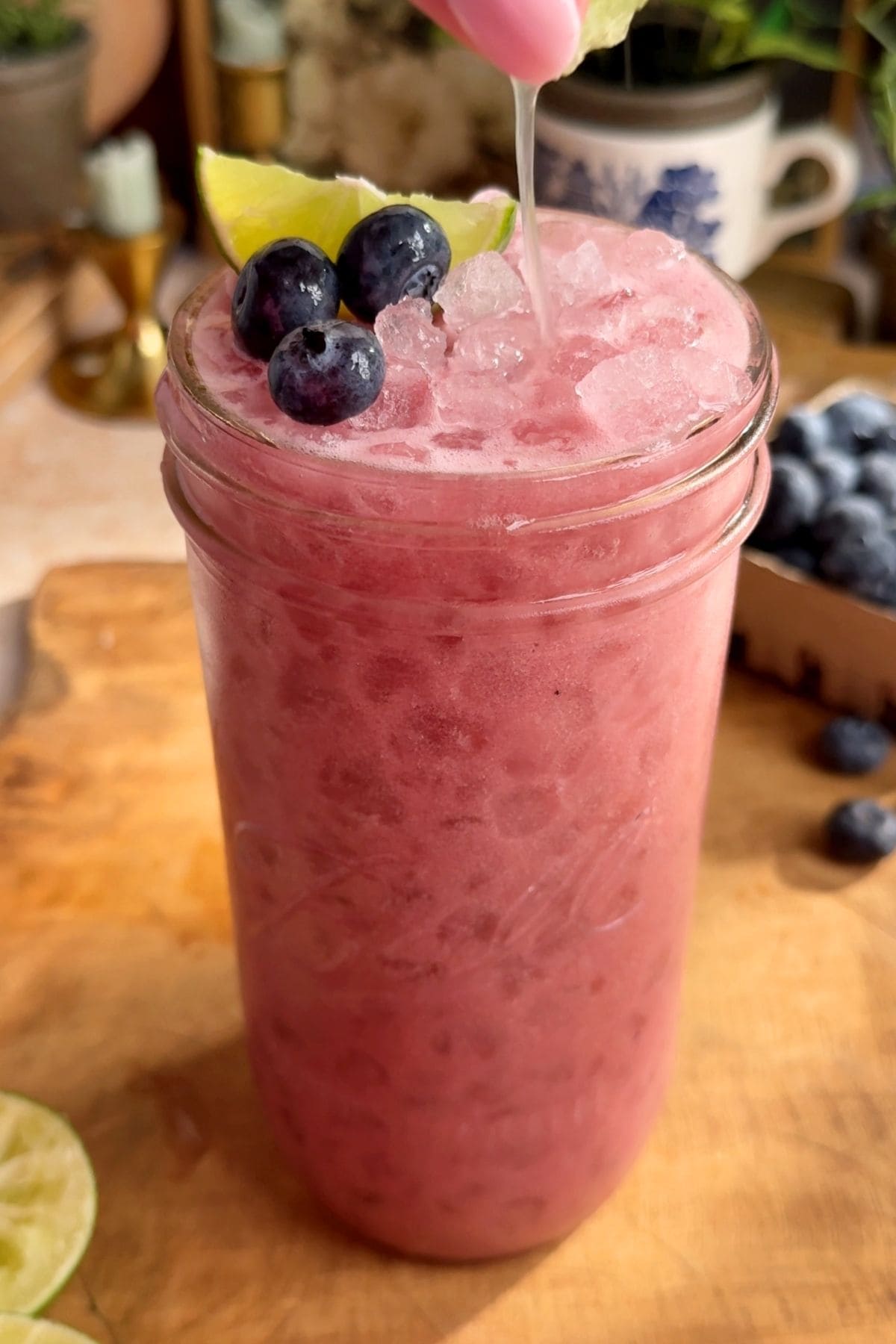 A glass of homemade blueberry Gatorade with a deep violet hue, served over ice and garnished with fresh blueberries and lime slices on a wooden surface with decorative plants, candles, and blueberries behind it. A visible thumb is seen squeezing fresh lime juice into the glass.