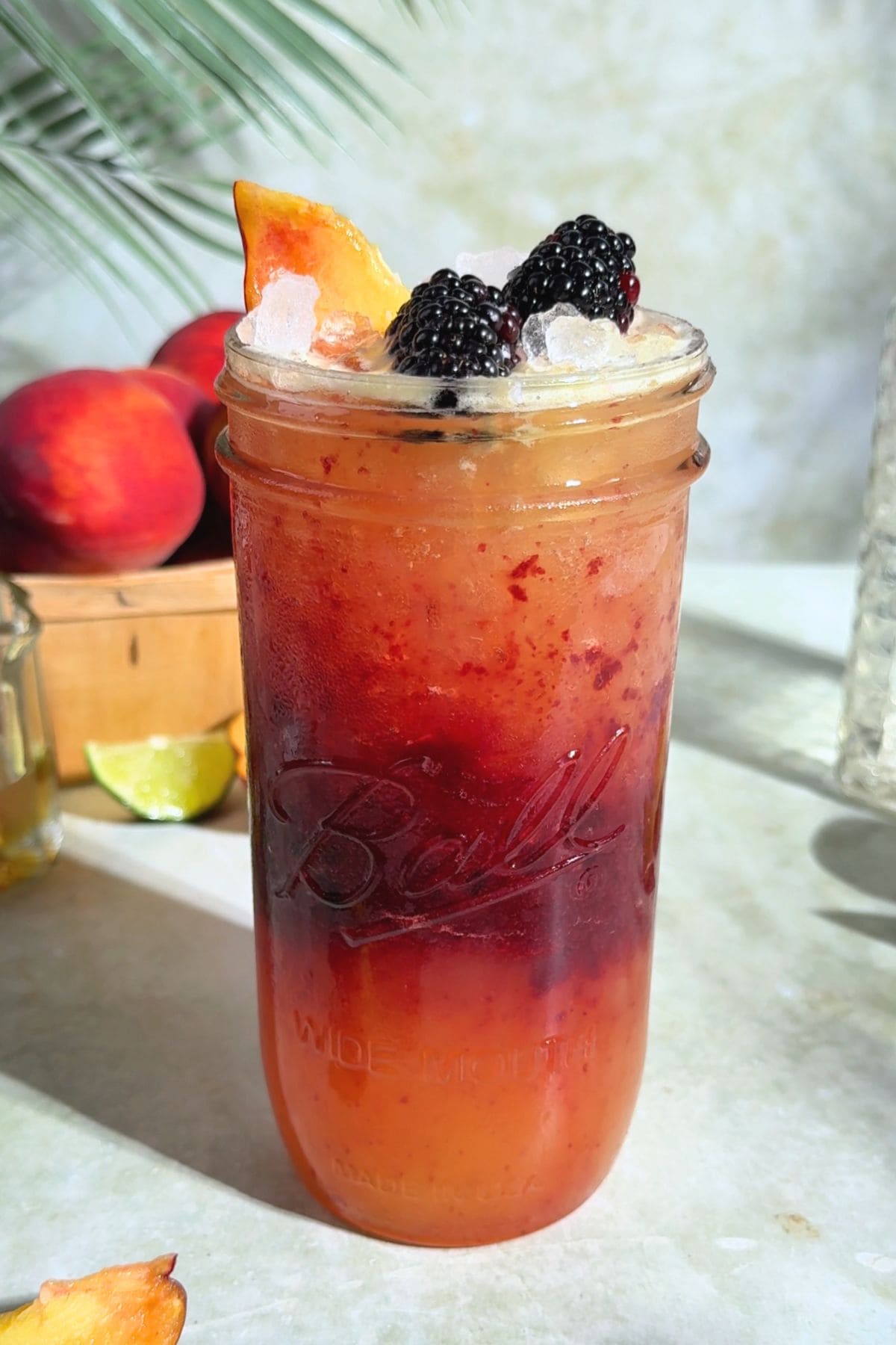 A glass of homemade blackberry peach Gatorade with a peach-toned top layer and dark blackberry purée in the middle, served over ice and garnished with a fresh peach slice and two blackberryies. Set on a light colored surface with soft natural light.