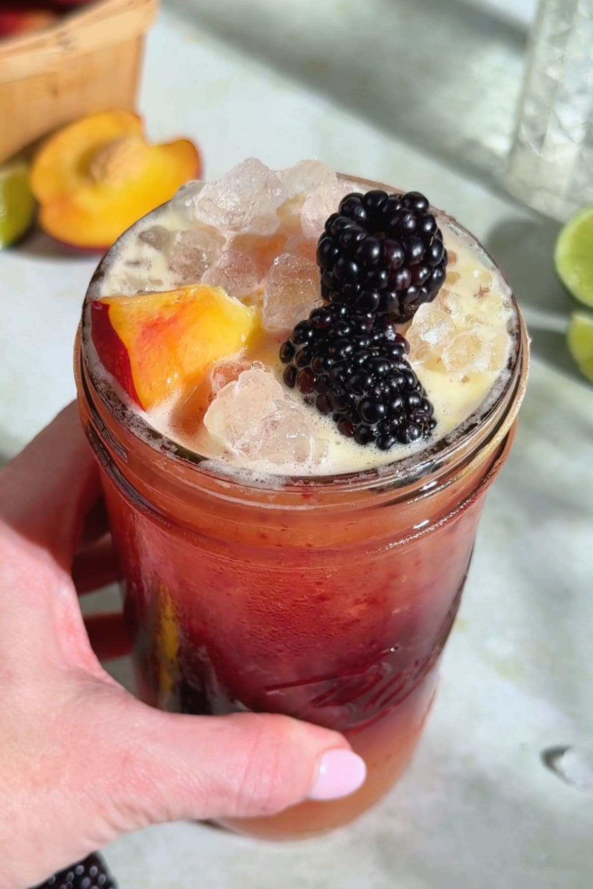 Overhead view of a hand holding a glass of blackberry peach Gatorade, showing distinct layered colors from the fruit purée and peach blend. Garnished with fresh blackberries and peach slices, surrounded by scattered fruit on a light backdrop.
