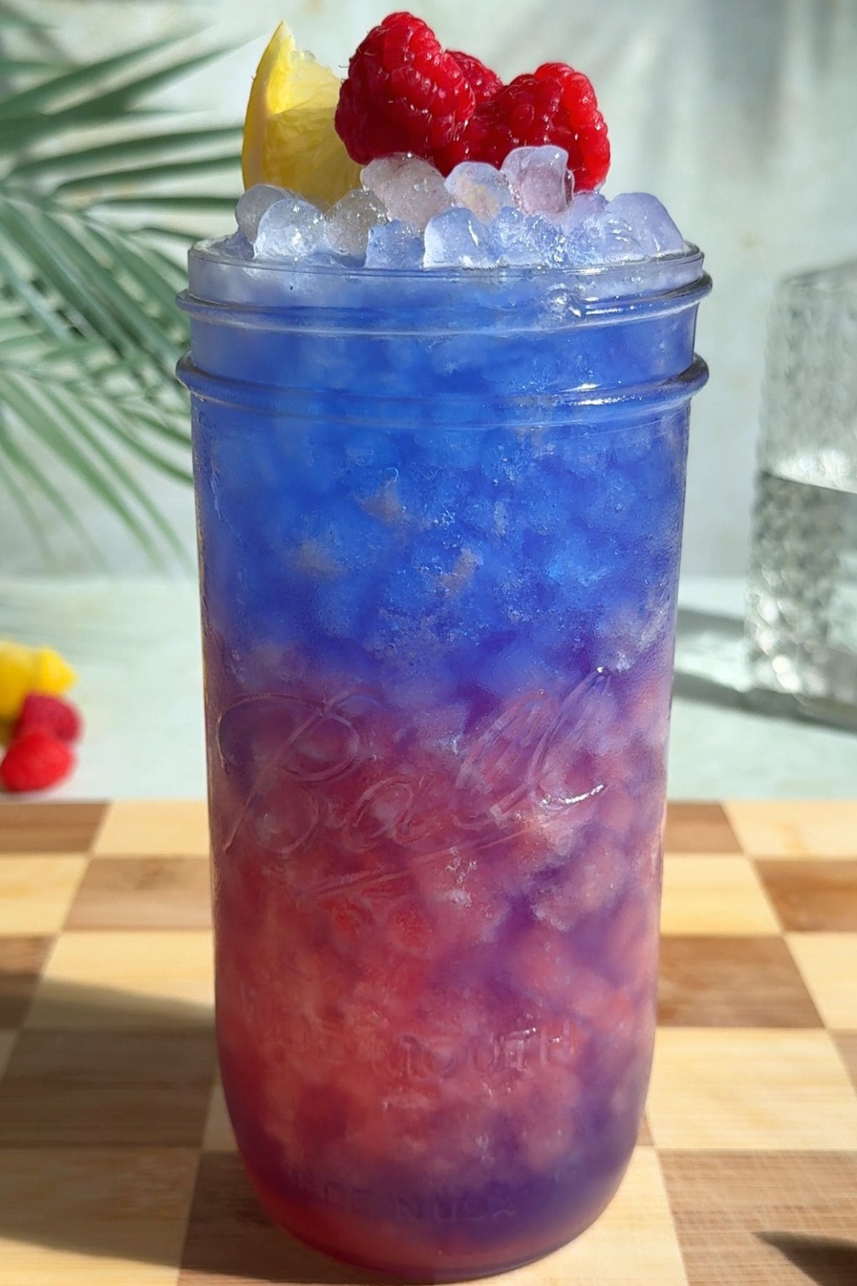 A tall glass of vibrant homemade blue raspberry Gatorade with a layered pink and electric blue hue, served over ice and garnished with a lemon wedge and fresh raspberries on a wooden surface.