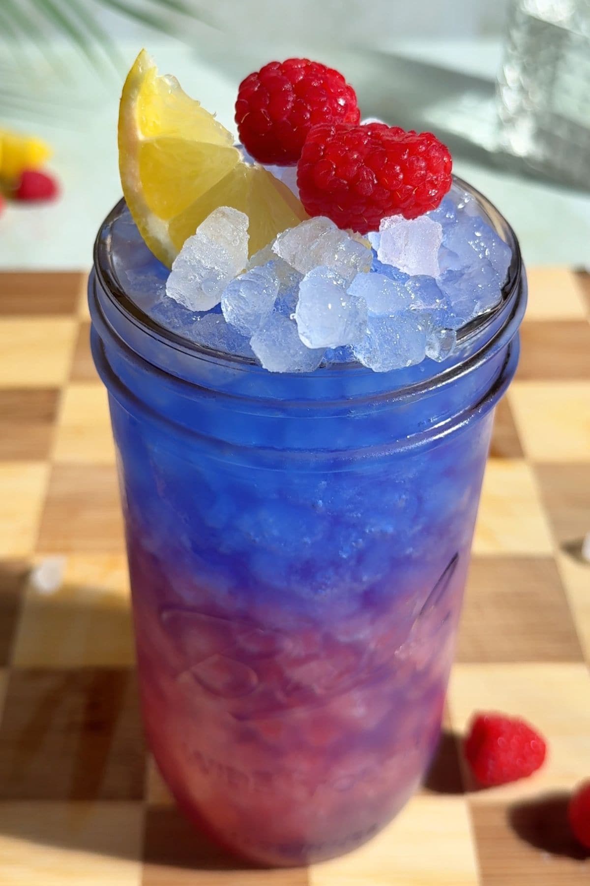 A close up of homemade blue raspberry Gatorade with a layered pink and electric blue hue, served over ice and garnished with a lemon wedge and fresh raspberries on a wooden surface with a couple of raspberries lying next to the glass.