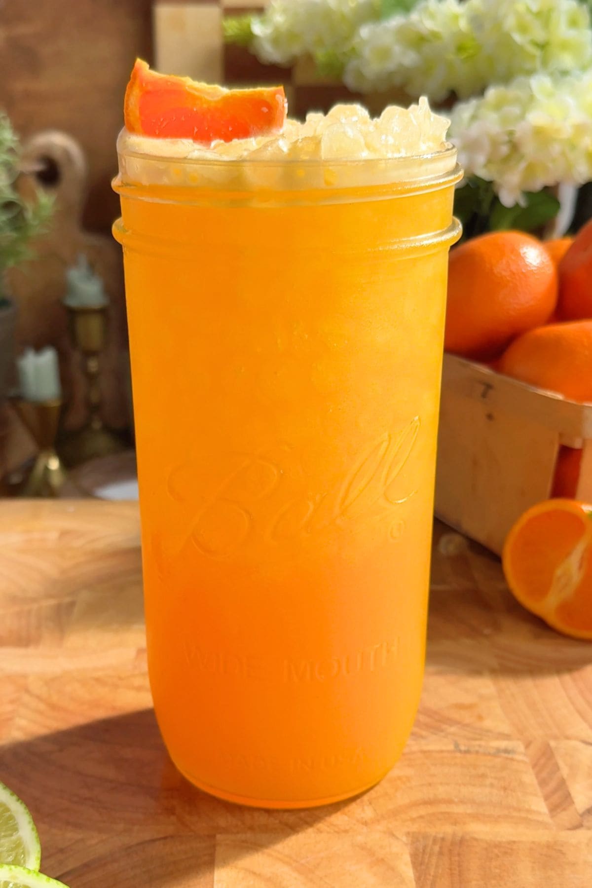 A glass of homemade orange Gatorade-inspired drink with a vibrant citrus color, served over ice and garnished with a cutie wedge. Set against a sunny kitchen backdrop with fresh oranges and limes scattered nearby.