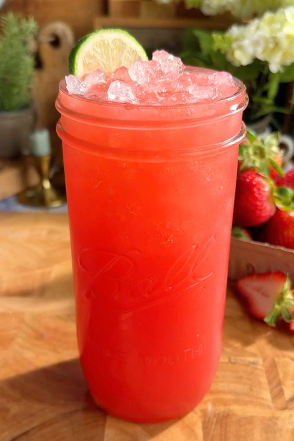 A glass of homemade strawberry Gatorade with a rosy pink hue, served over ice and garnished with a lime wedge. Fresh strawberries are scattered on a wooden surface behind the glass.