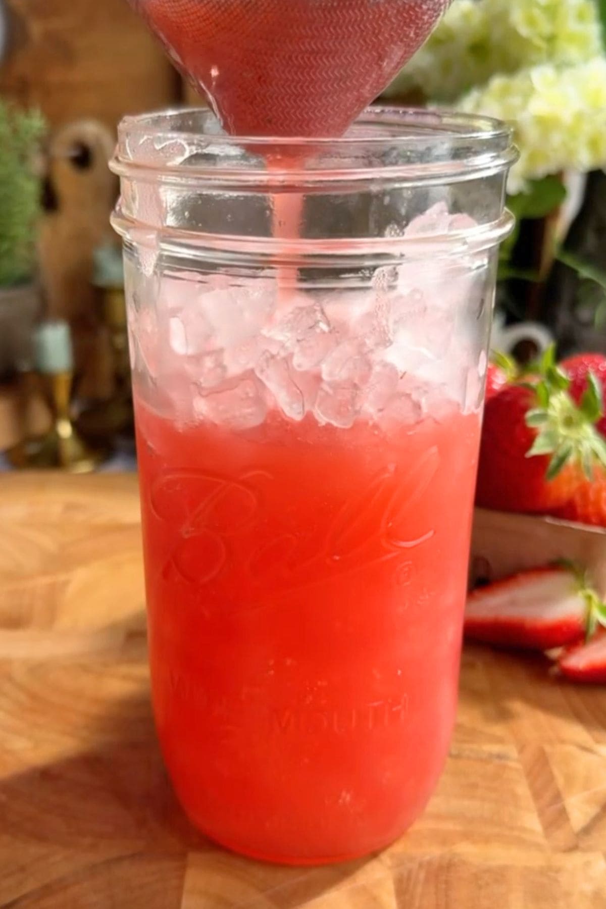 A glass half full of homemade strawberry Gatorade with a sieve straining more into the glass, served over ice. Fresh strawberries are scattered on a wooden surface behind the glass.