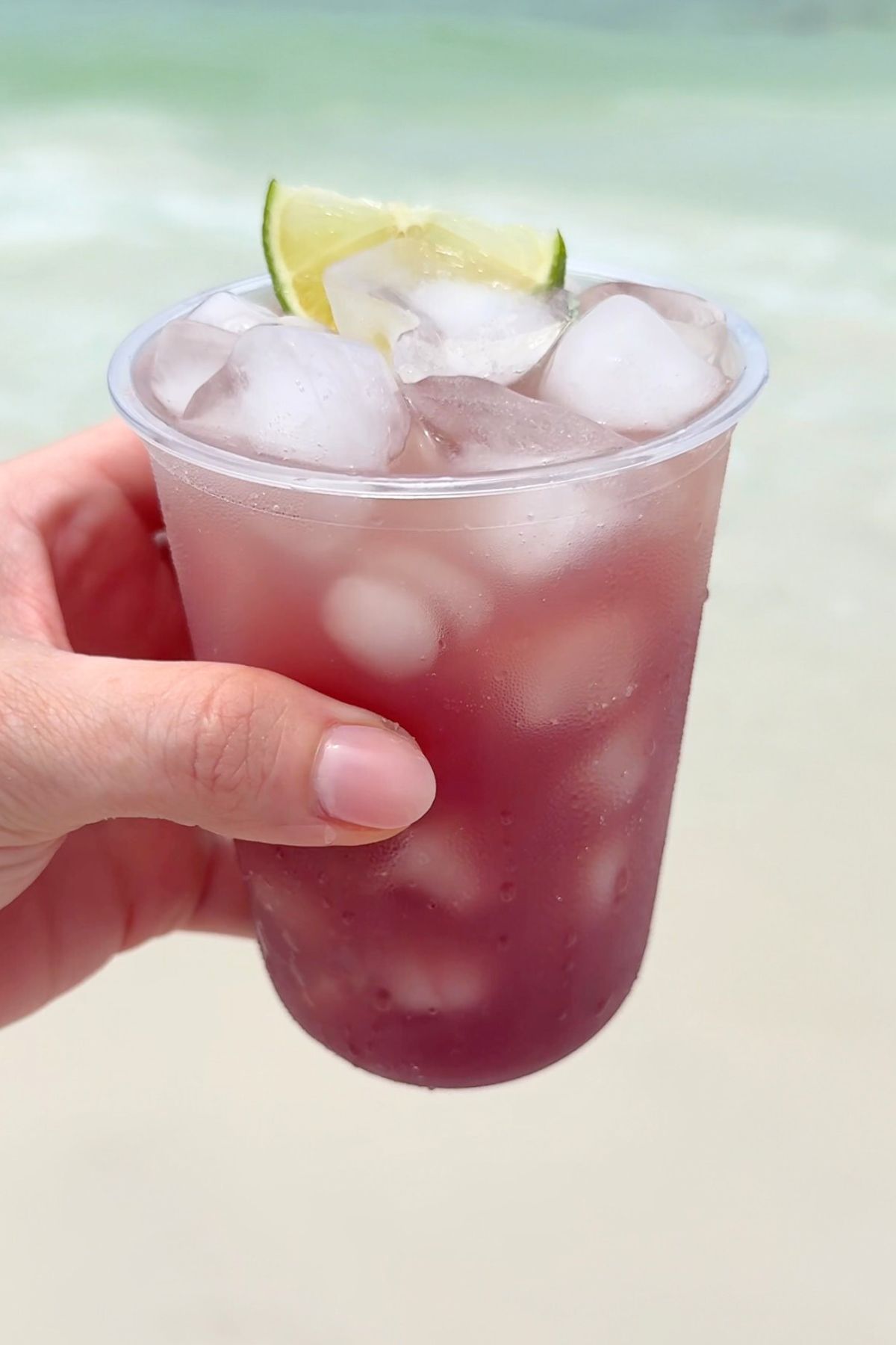 A hand holding a clear plastic cup of tropical beach Gatorade served over ice with a lime wedge set in front of the ocean.