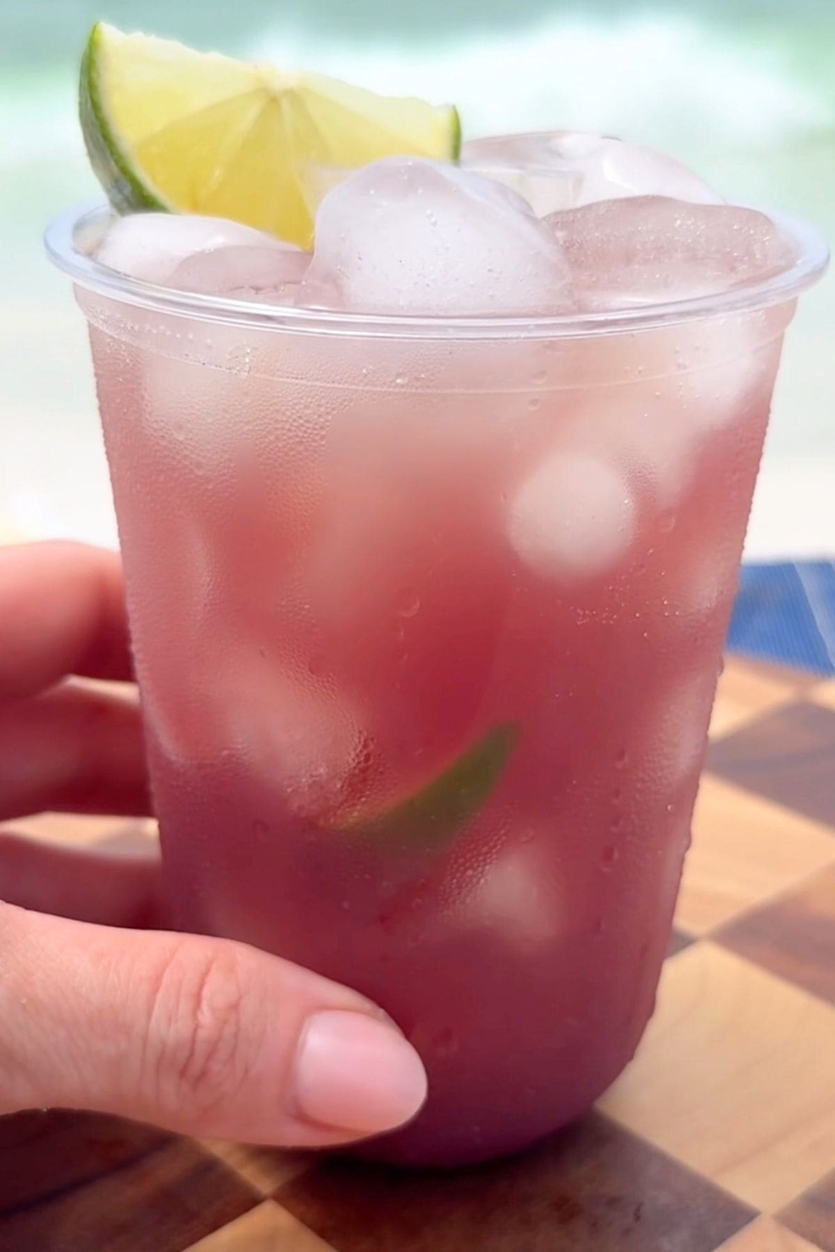 A close-up of a hand holding a clear cup of tropical beach Gatorade served over ice with a lime wedge on a checkerboard wood surface.