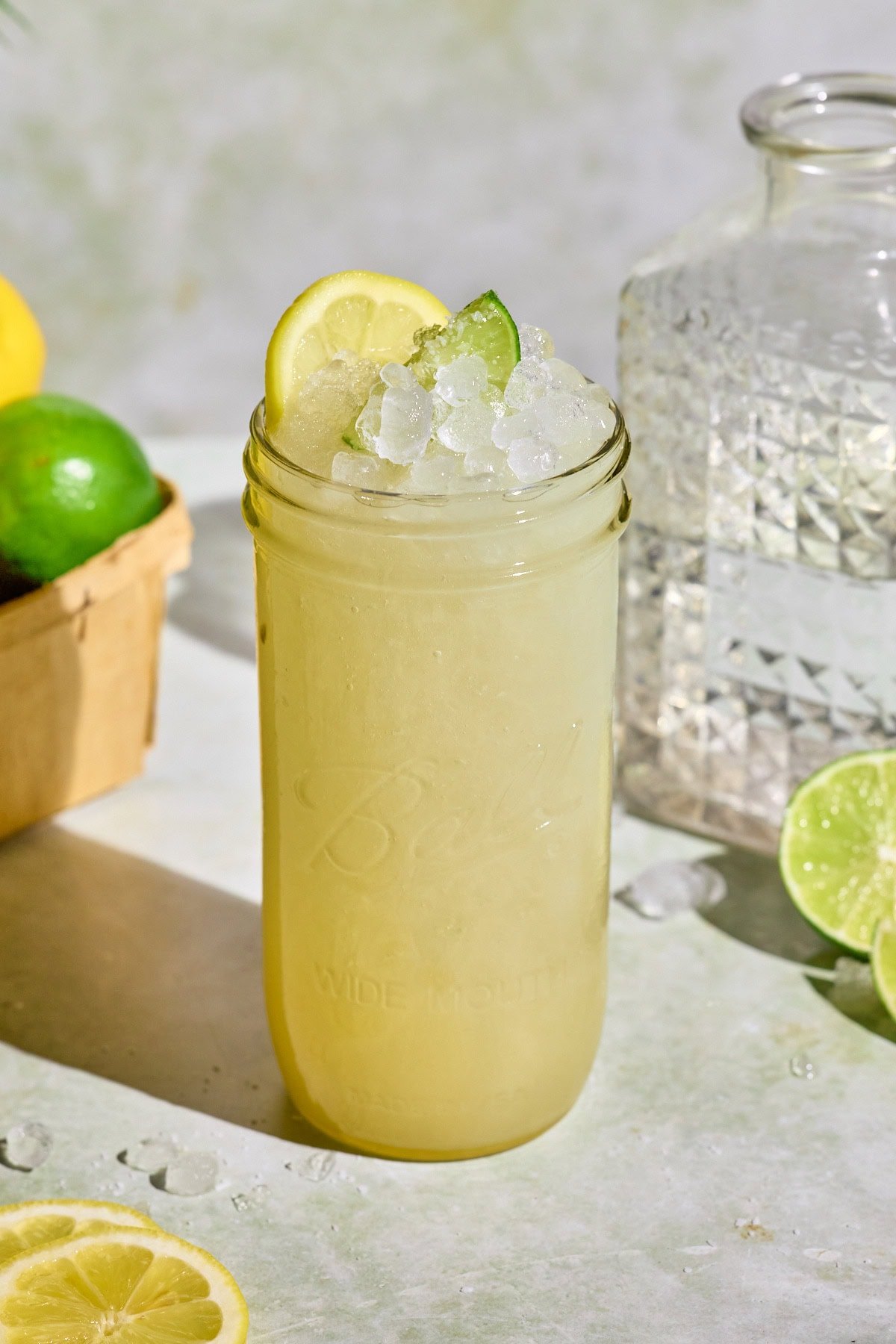 A chilled glass of homemade lemon lime Gatorade served over ice and garnished with fresh citrus slices. Set on a light colored sunlit surface alongside a glass pitcher, halved limes, and a woven basket brimming with lemons and limes.