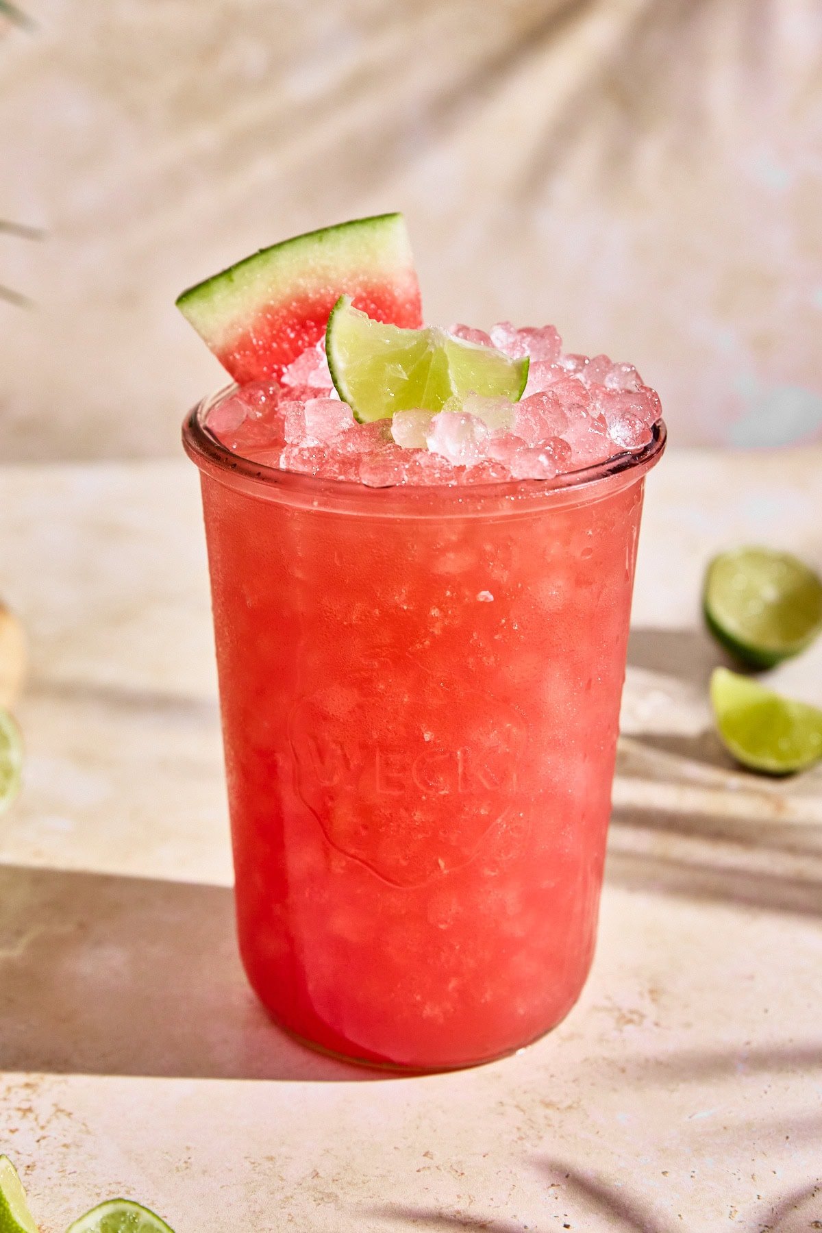 A glass of homemade watermelon limeade Gatorade with a soft pink hue, served over ice and garnished with a fresh lime wedge and a slice of watermelon. The glass is placed on a light-colored surface with lime slices in the background.