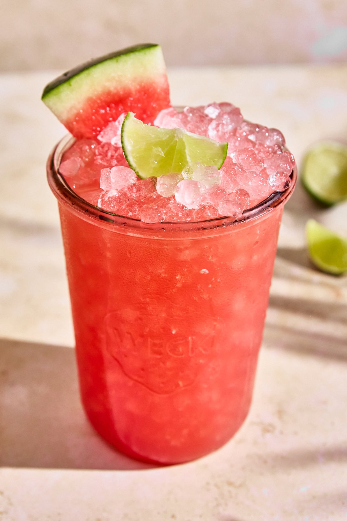 Overhead view of a glass of watermelon limeade Gatorade served with pebble ice,  a lime wedge and a small watermelon slice. Set on a light backdrop with a halved lime in the background.