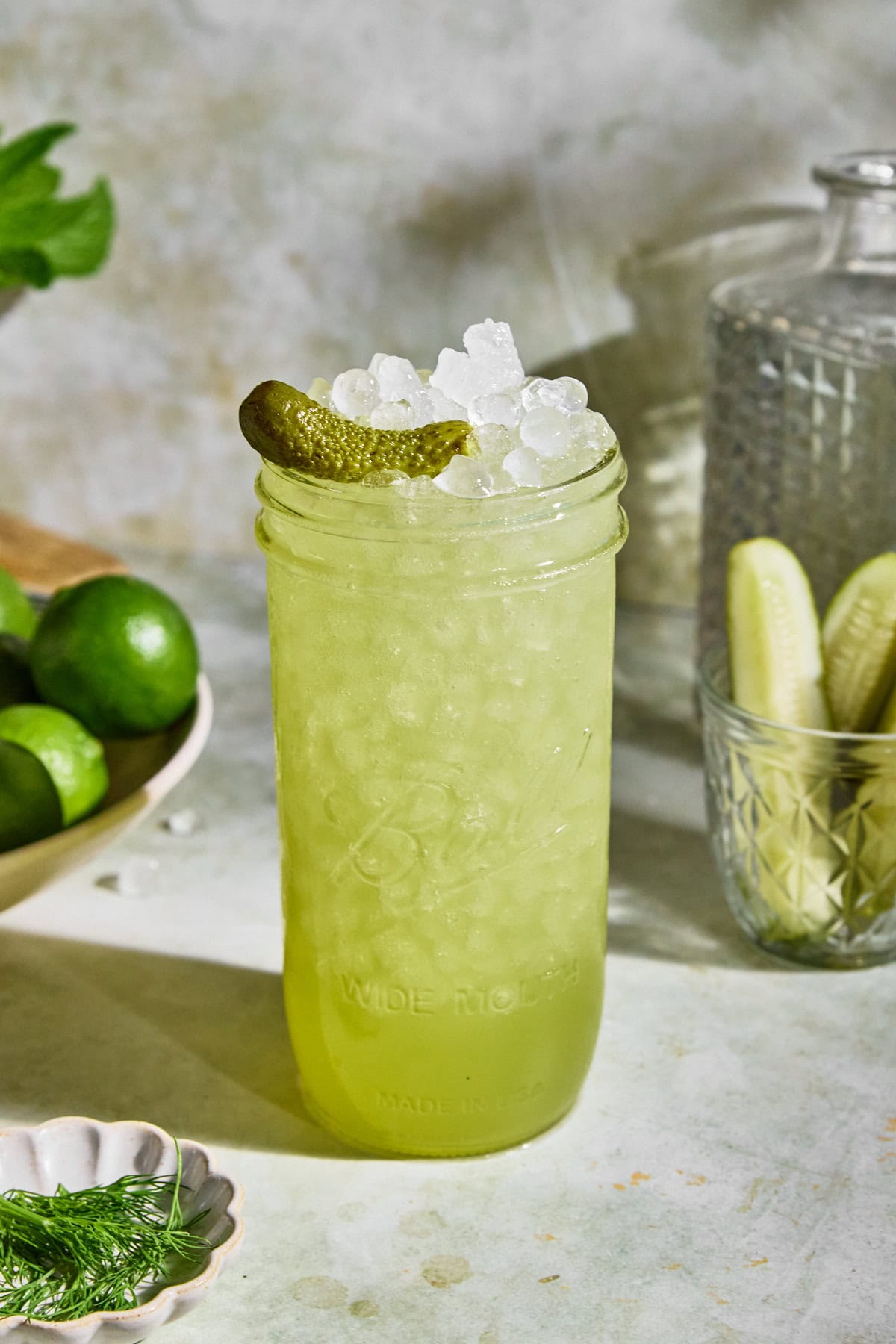 A chilled glass of dill pickle electrolyte drink on a light surface with a light green hue, garnished with a pickle spear. Behind the glass is a jar of more pickle spears and a bowl of fresh limes.