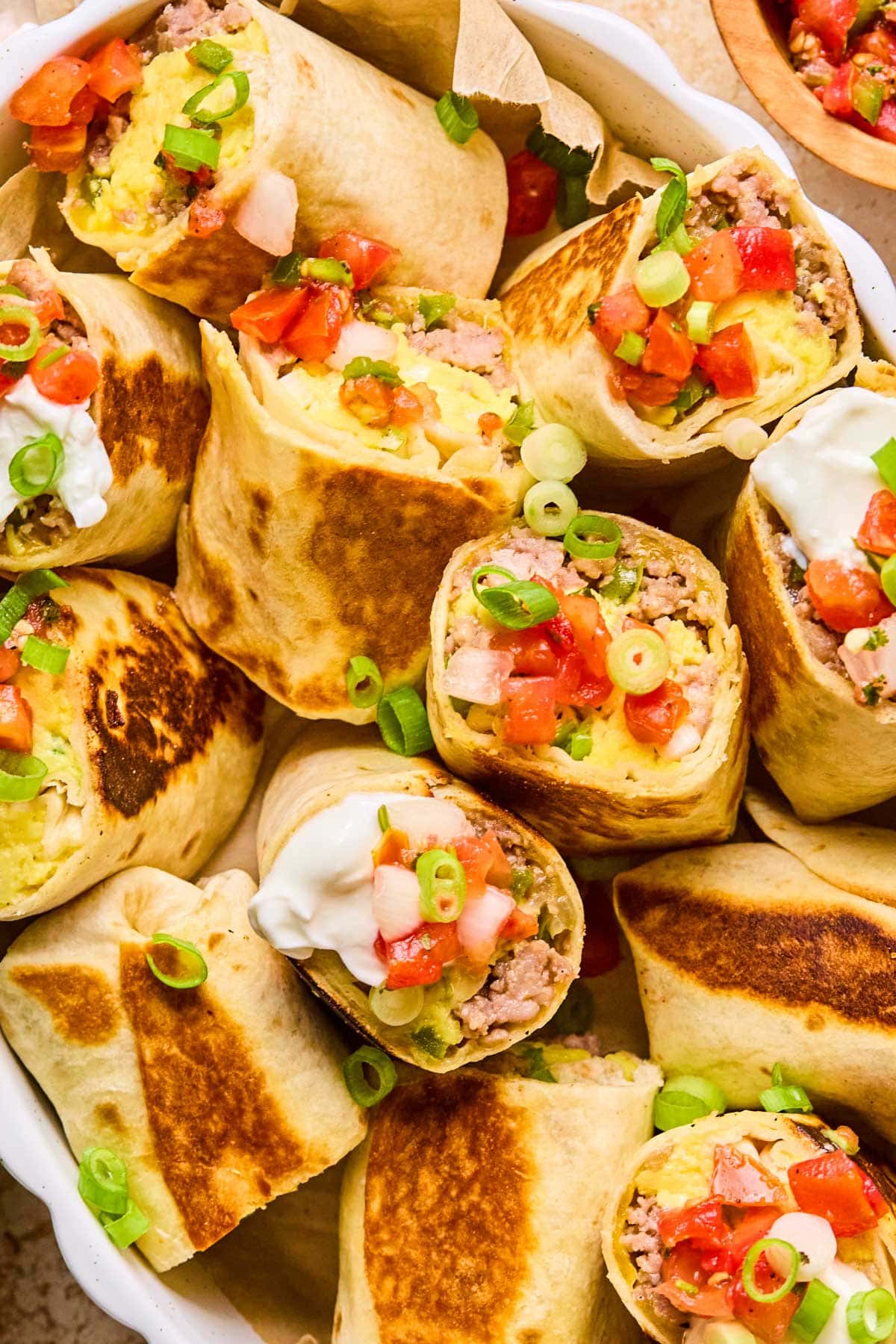 Close-up of halved breakfast burritos in a white ceramic dish, ready to freeze or eat. Garnished with green onions and tomato slices.