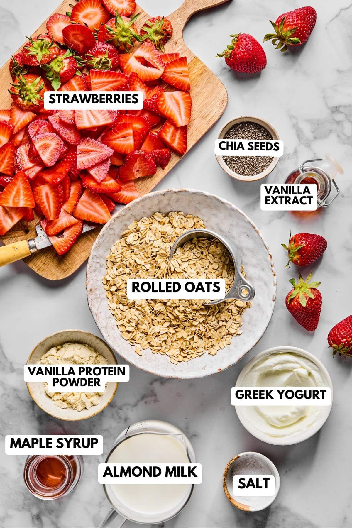 Flat lay of ingredients for high protein overnight oats including oats, strawberries, Greek yogurt, almond milk, chia seeds, and protein powder on a light gray and white countertop.