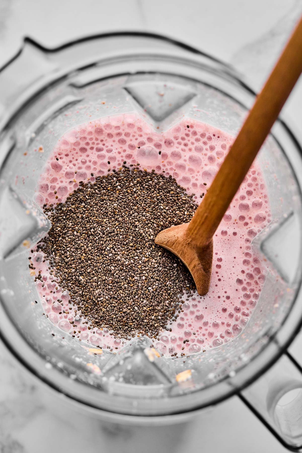 A blender with blended strawberries, oats, Greek yogurt, almond milk, and protein powder, topped with chia seeds before mixing them in.