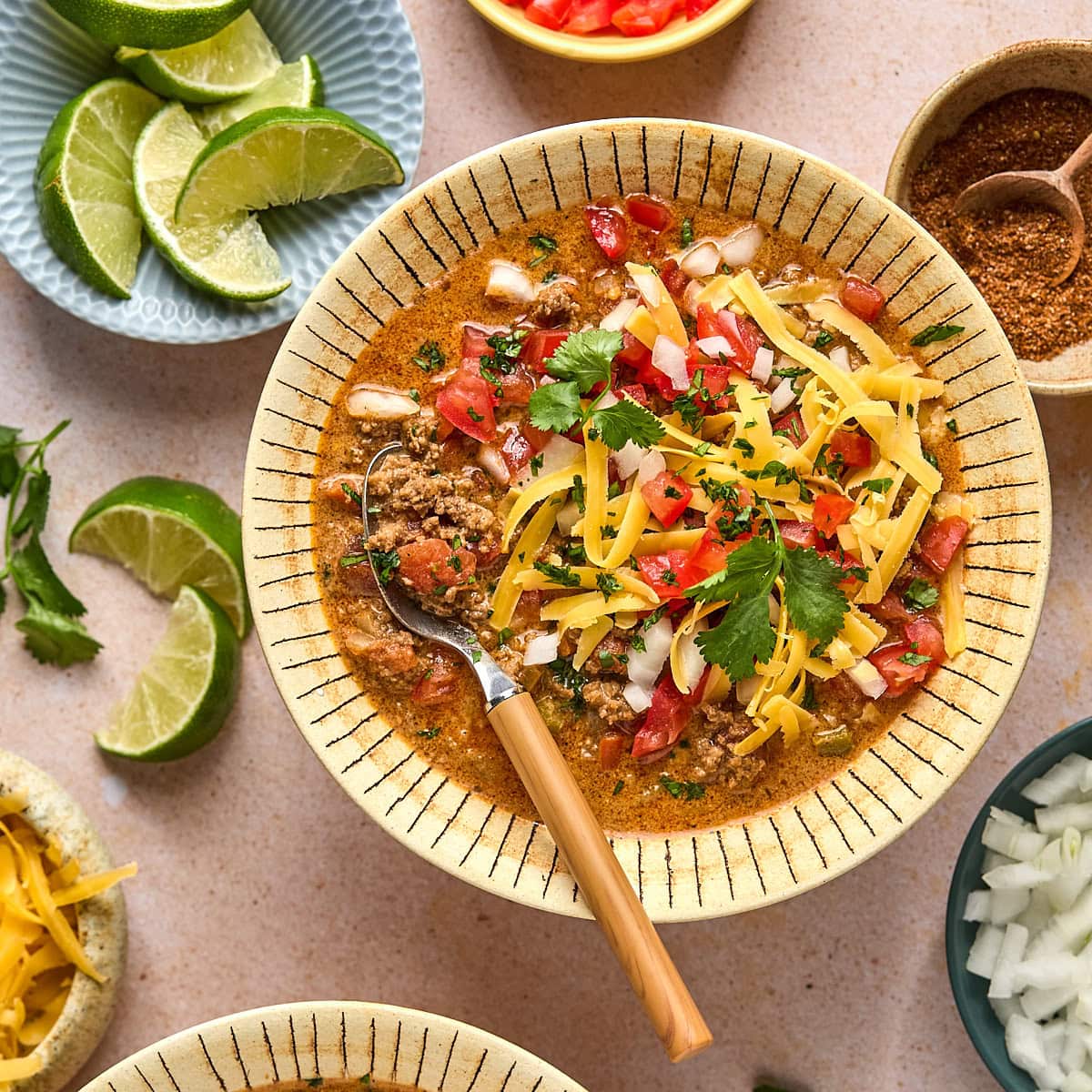Bowl of creamy taco soup topped with shredded cheese and fresh cilantro, served with a spoon and surrounded by little bowls with limes, onions, and spices on a light pink backdrop.