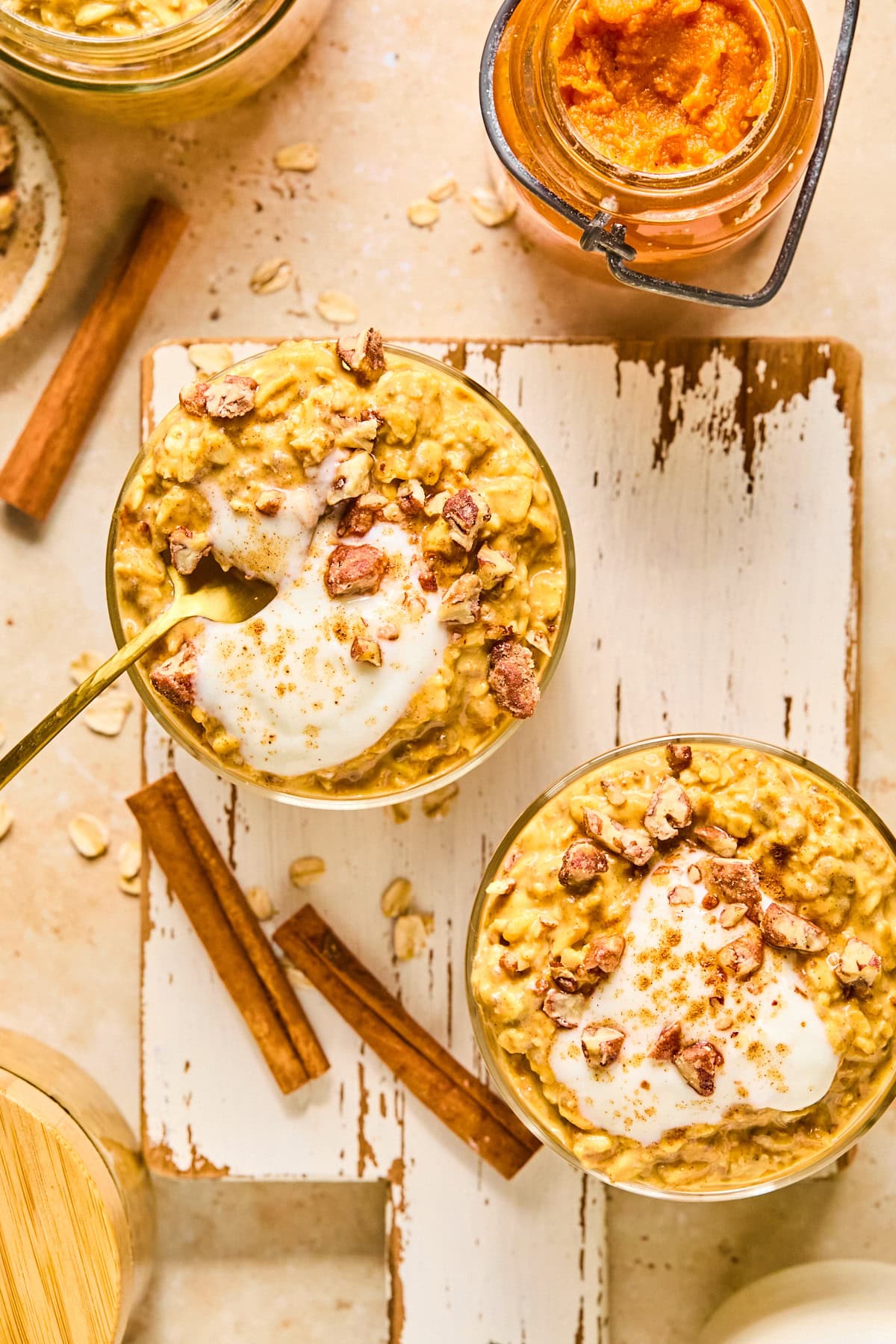 Overhead-shot of pumpkin pie overnight oats topped with maple-sweetened Greek yogurt and chopped honey roasted pecans on a wooden board, with a pink backdrop and fall baking ingredients in the background.