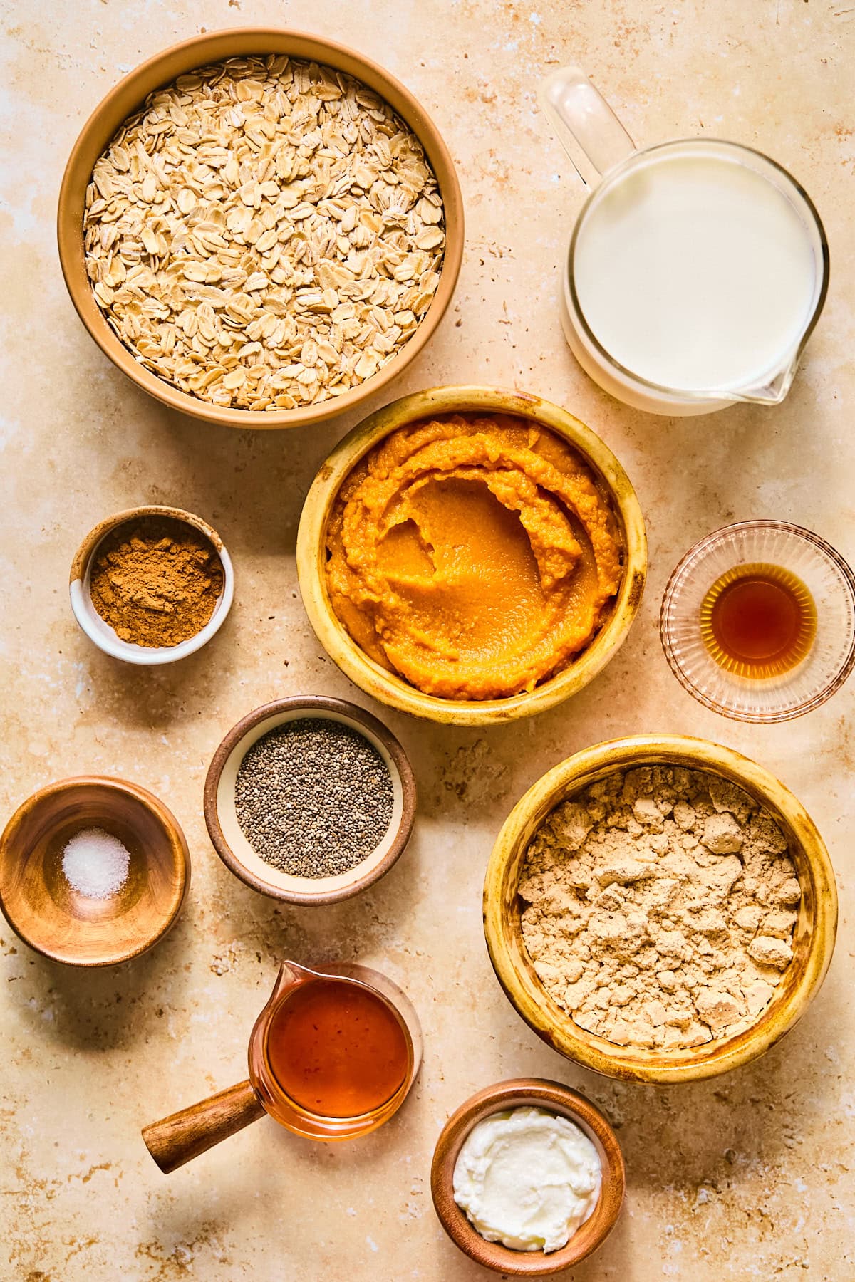Ingredients for pumpkin pie overnight oats on a light colored counter in decorative bowls including oats, unsweetened almond milk, pumpkin puree, greek yogurt, vanilla protein powder, chia seeds, pure maple syrup, pumpkin pie spice, and vanilla extract.