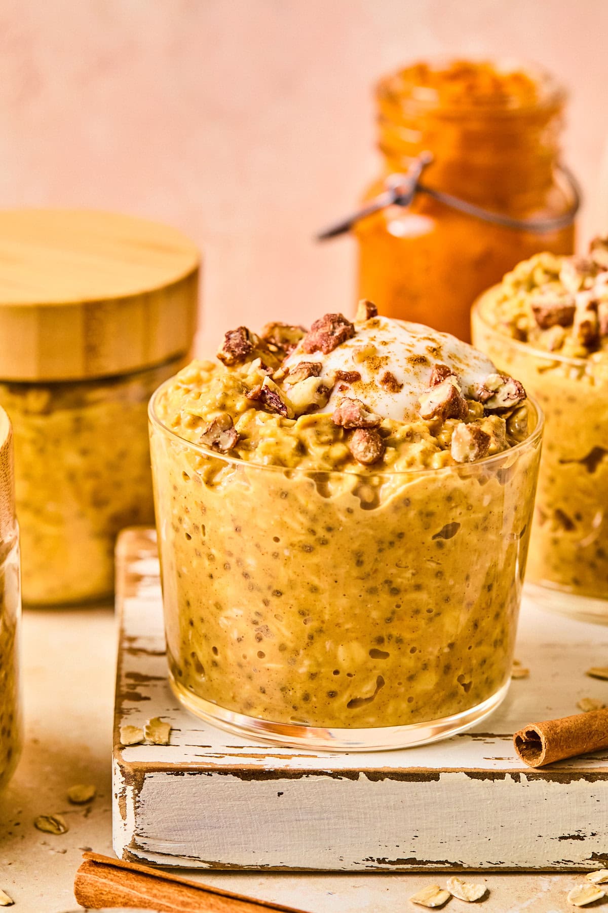 Close-up of pumpkin pie overnight oats in glass jars topped with maple-sweetened Greek yogurt and chopped honey roasted pecans on a wooden board, with a pink backdrop and fall baking ingredients in the background.
