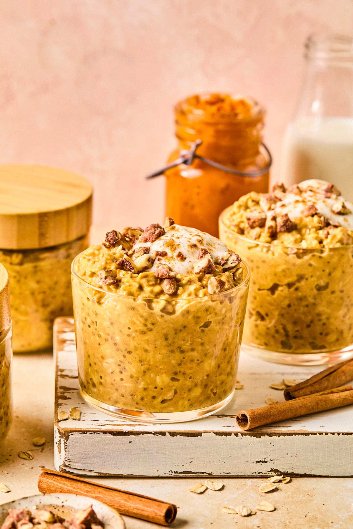 Glass jars filled with pumpkin pie overnight oats topped with maple-sweetened Greek yogurt and chopped honey roasted pecans on a wooden board, with a pink backdrop and fall baking ingredients in the background.