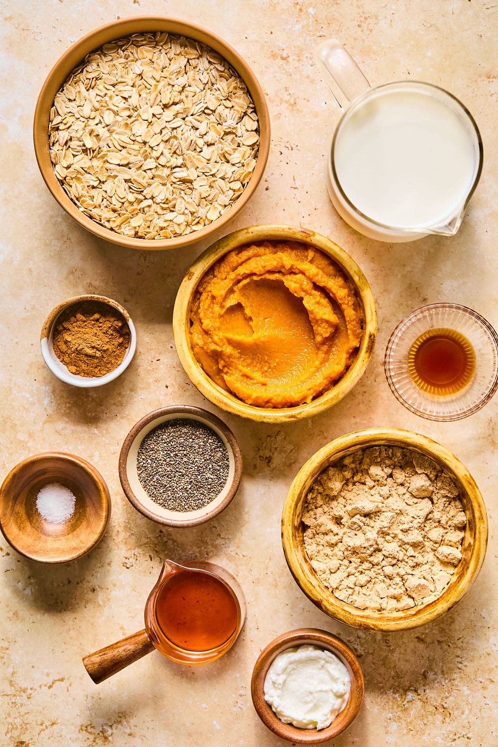 Ingredients for pumpkin pie overnight oats on a light colored counter in decorative bowls including oats, unsweetened almond milk, pumpkin puree, greek yogurt, vanilla protein powder, chia seeds, pure maple syrup, pumpkin pie spice, and vanilla extract.
