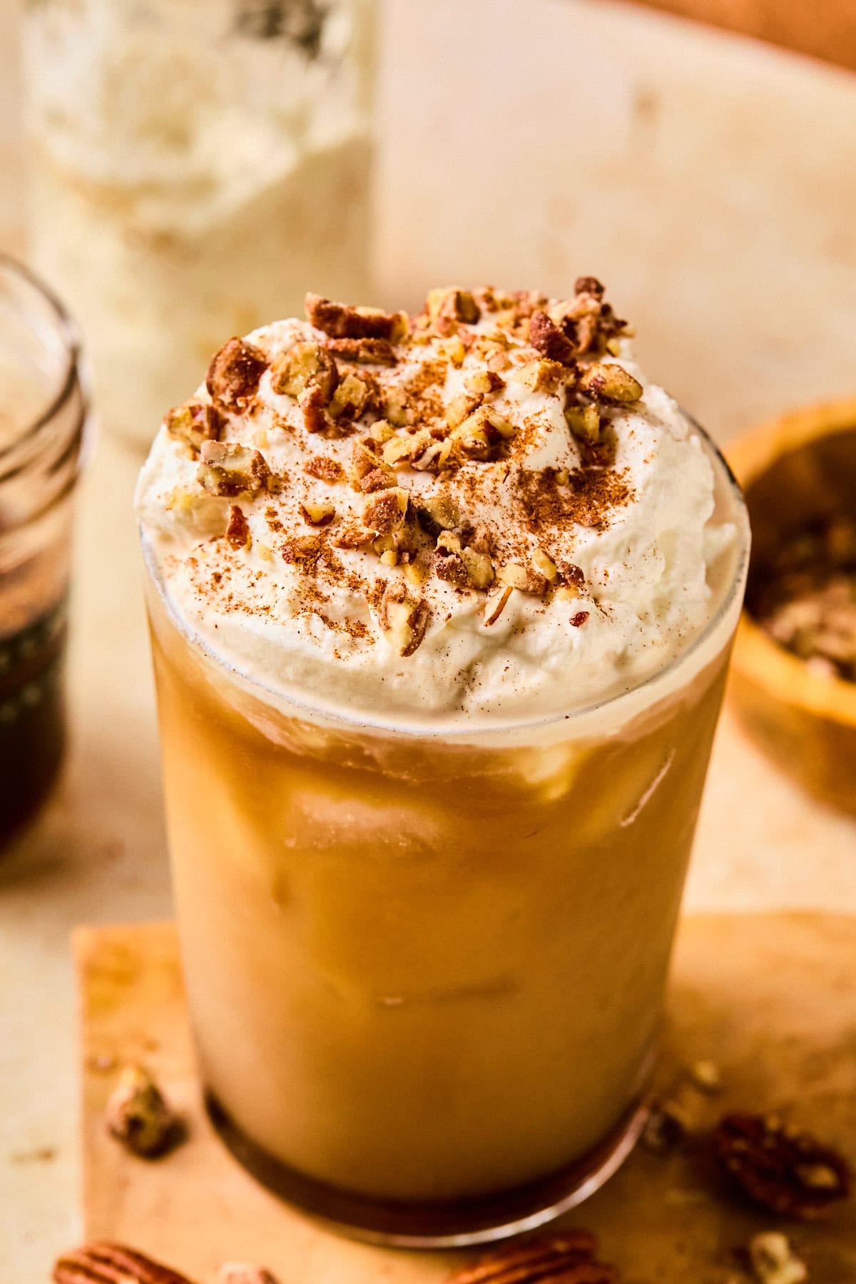 Close-up of an iced pecan crunch oatmilk latte in a tall glass topped with whipped oat cream, cinnamon, and chopped pecans.
