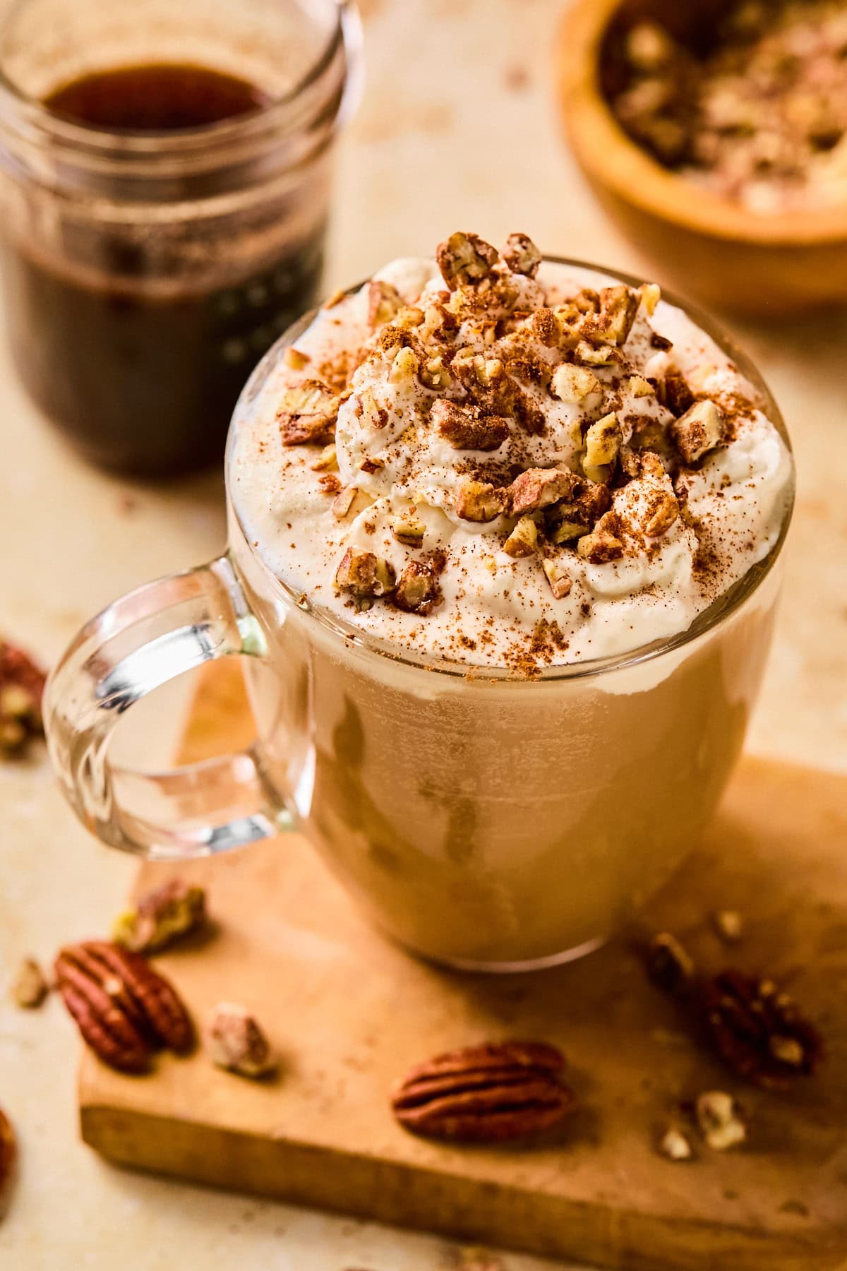Pecan crunch oatmilk latte in a glass mug topped with whipped oat cream, a drizzle of syrup, a sprinkle of cinnamon, and chopped pecans.