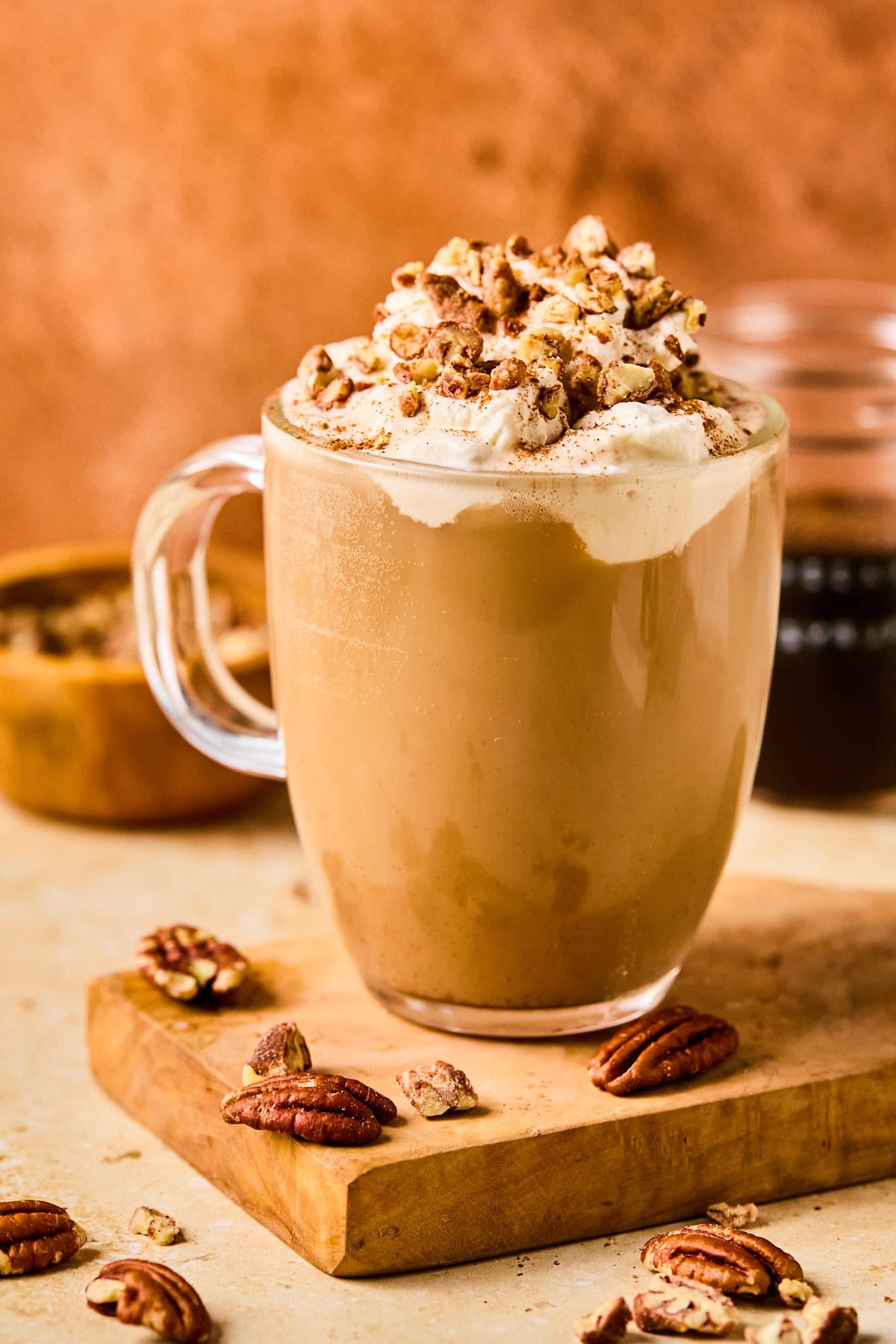 Pecan crunch oatmilk latte in a glass mug topped with whipped oat cream, a drizzle of syrup, a sprinkle of cinnamon, and chopped pecans.