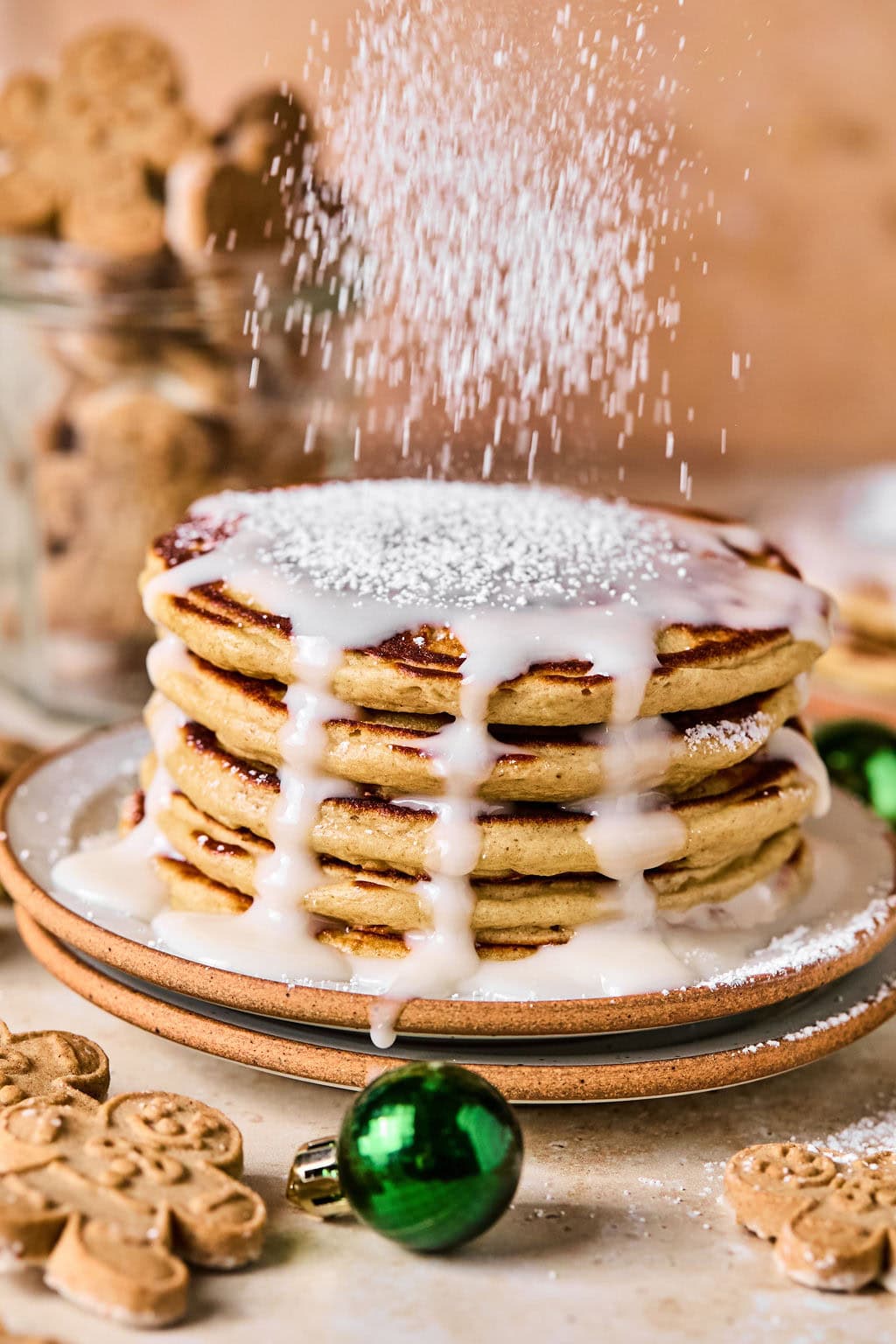owdered sugar being sprinkled over a stack of gingerbread pancakes topped with glaze, surrounded by gingerbread cookies and Christmas ornaments.