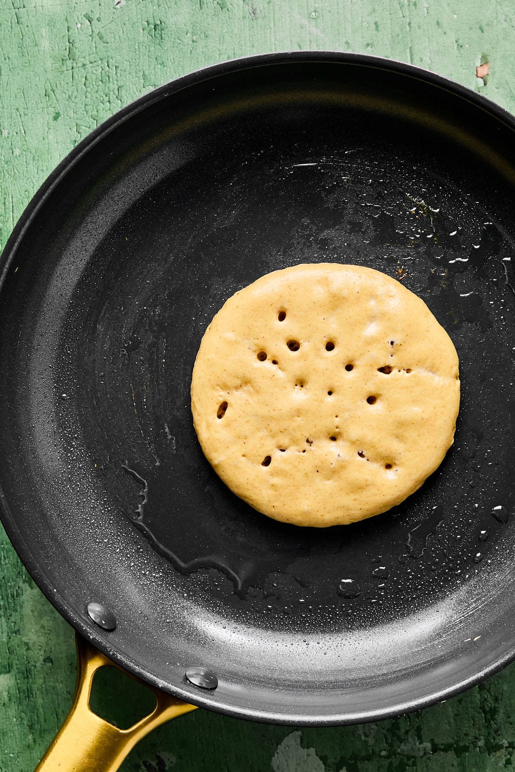 A 1/4 cup portion of gingerbread pancake batter cooking on a black griddle skillet, with small bubbles forming.