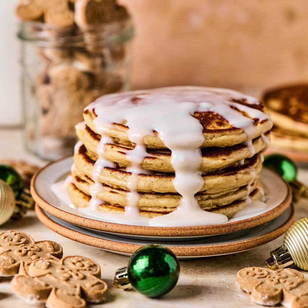 A tall stack of gingerbread pancakes drizzled with glaze on a plate, surrounded by gingerbread cookies and Christmas ornaments on a light backdrop.