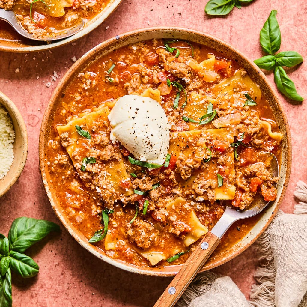 Bowl of lasagna soup topped with ricotta cheese and fresh basil, set on a pink countertop with extra basil leaves and a small bowl of parmesan to the side.
