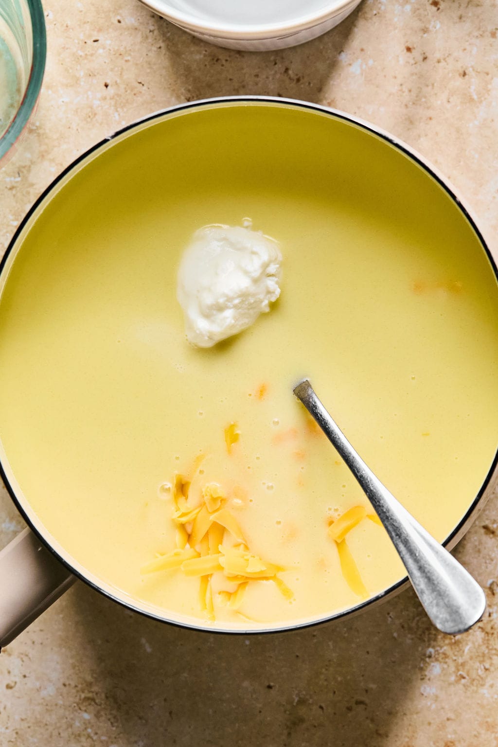 Cheddar and Greek yogurt added to the small saucepan of sauce with a spoon resting inside.