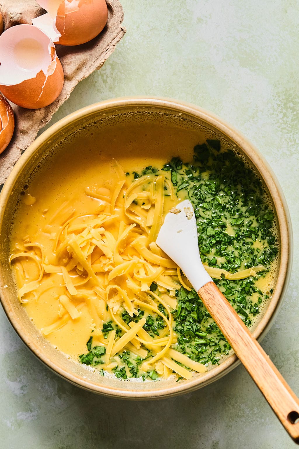 Eggs and cottage cheese whisked in a large bowl with cheddar cheese and spinach on top, and a spatula resting against the bowl ready to fold everything together.