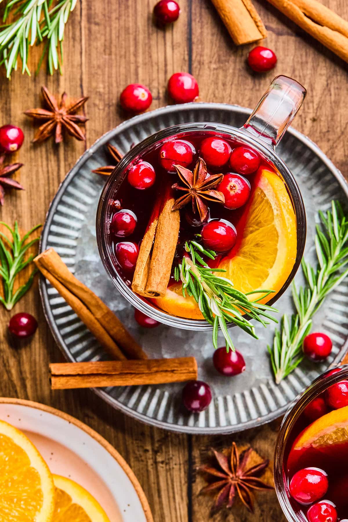Overhead view of non-alcoholic mulled wine in a glass mug on a silver serving plate, garnished with an orange slice, cinnamon sticks, whole cloves, star anise, and a sprig of rosemary.